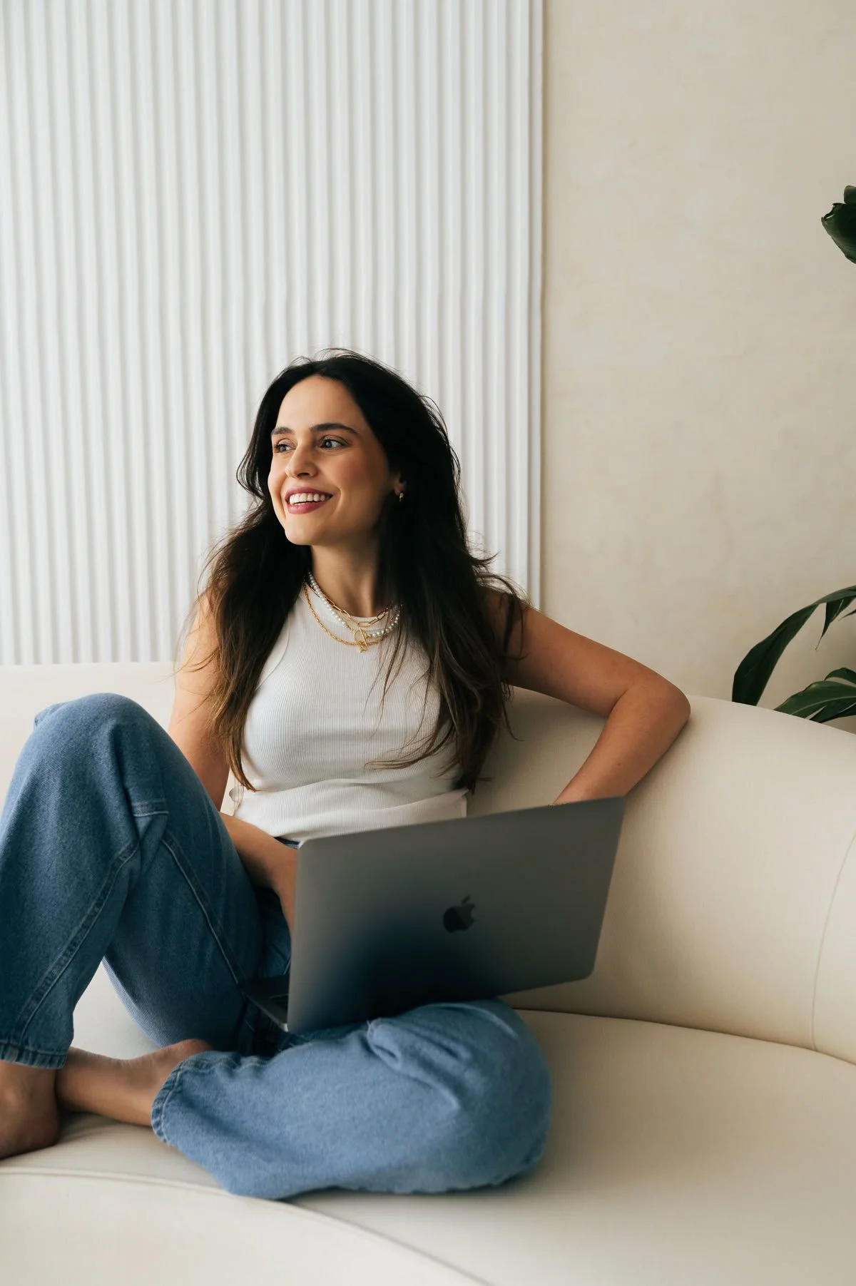 A woman sitting on a white couch with her legs crossed, smiling, while using a silver MacBook laptop. She is wearing a sleeveless white top, blue jeans, and layered gold necklaces. There is a green plant in the background near a wall with vertical blinds.