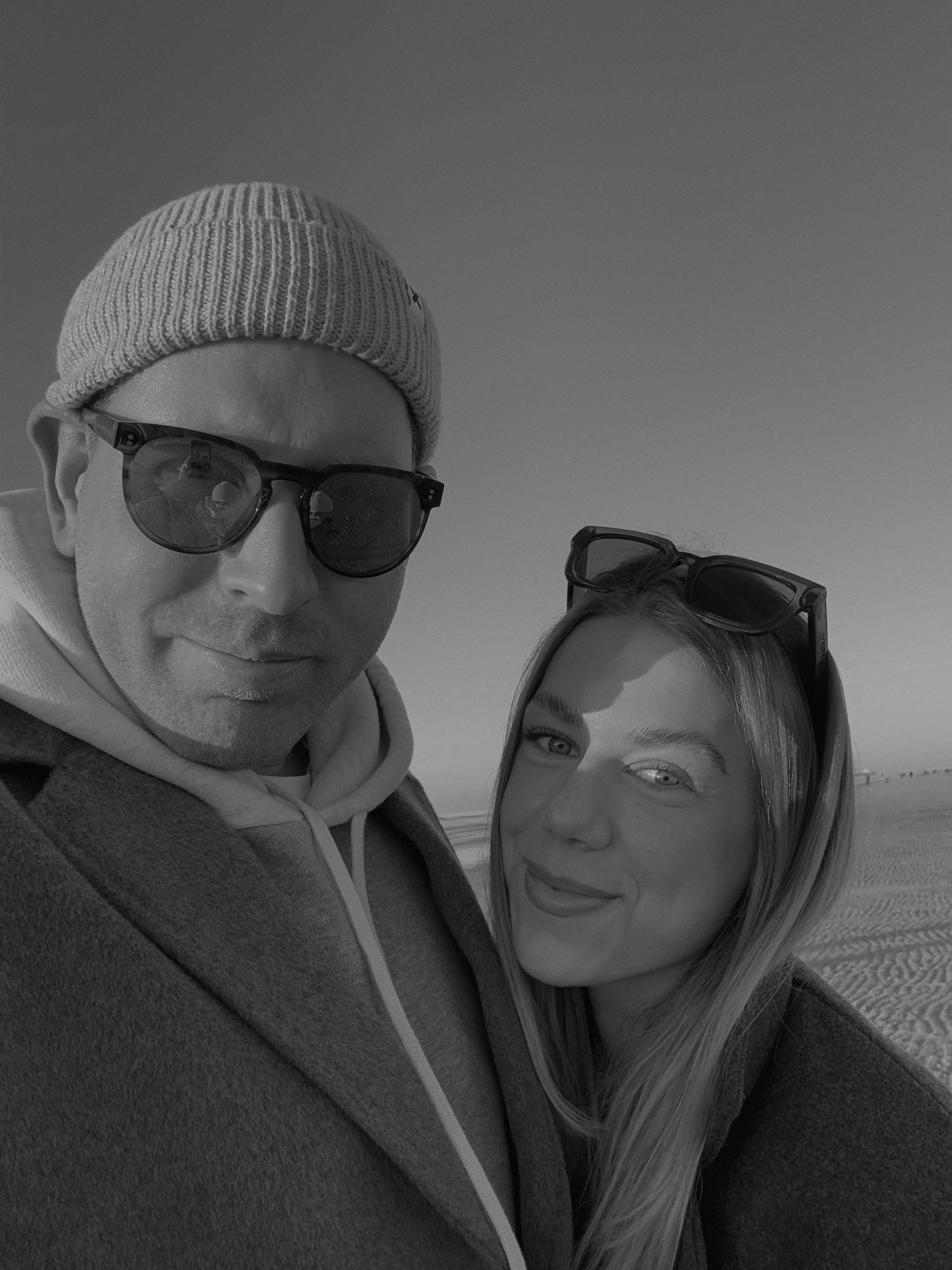 Ein liebevolles Paar macht ein Selfie im Freien bei Sonnenlicht, beide tragen Sonnenbrillen und lächeln, im Hintergrund ein Himmel und Sanddünen.