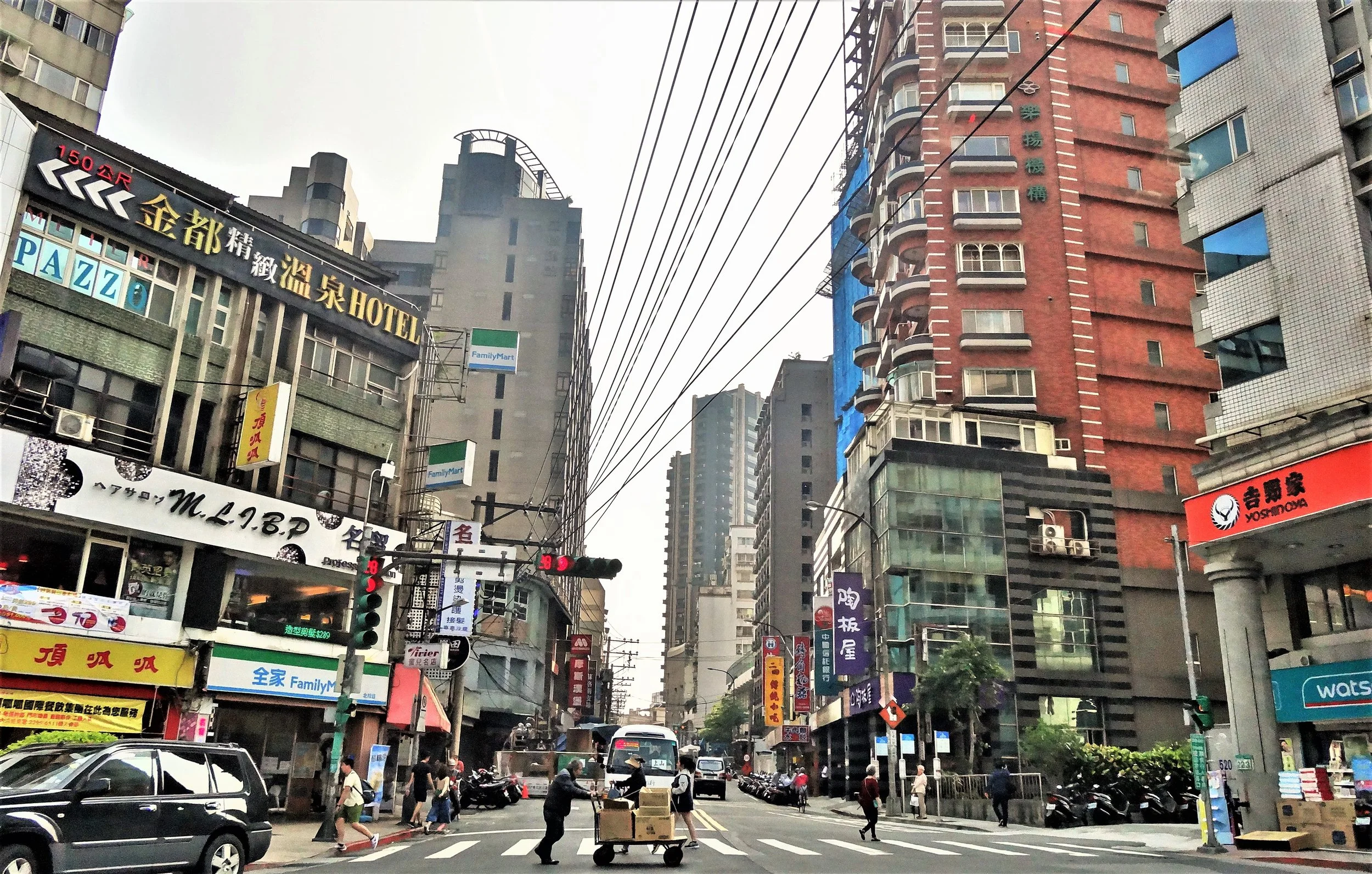  A busy city street with tall buildings, shops with colorful signs in Chinese, cars, and people crossing the road. Overhead, power lines crisscross the sky between the buildings. 