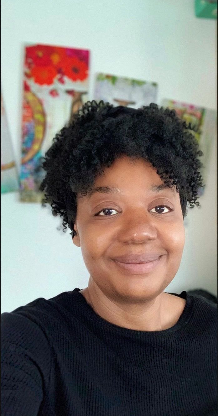 Retrato de una mujer con cabello rizado y sonrisa, fondo con posters coloridos en la pared.