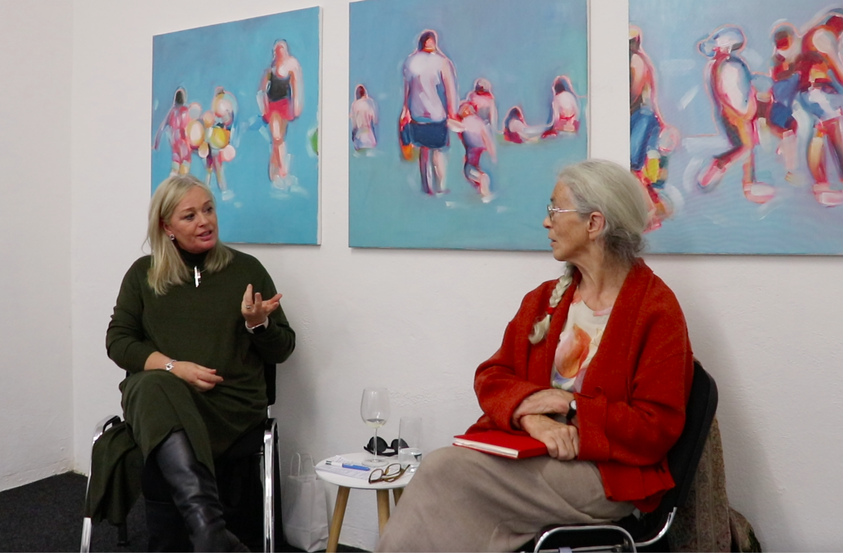Two women having a conversation in an art gallery. There are three colorful paintings of people at the beach hanging on the white wall behind them.