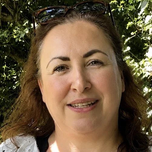 A woman with light brown hair wearing sunglasses on her head, smiling outdoors in front of green foliage.