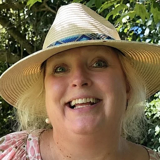 A woman smiling outdoors wearing a large straw hat with blue decorative accents and a pink patterned top.