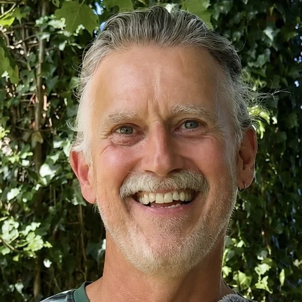 Smiling middle-aged man with gray hair and beard outdoors, lush green bushes in the background.