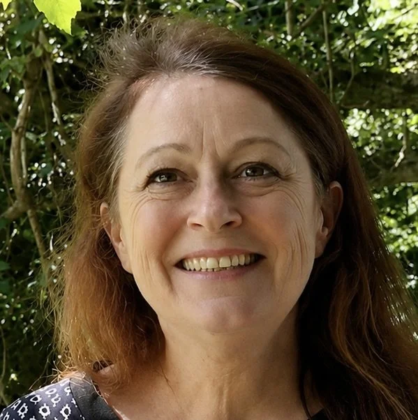 A woman smiling outdoors with green foliage in the background.