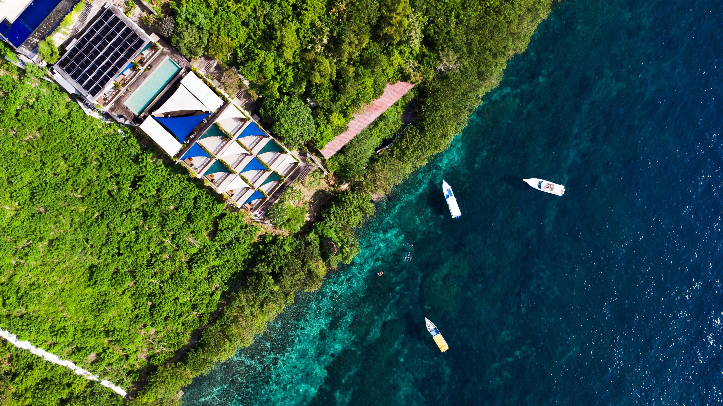 Vue aérienne d'une côte avec des appartements avec des toits en pente, entourés de verdure dense, et des bateaux flottant dans une mer bleue claire.