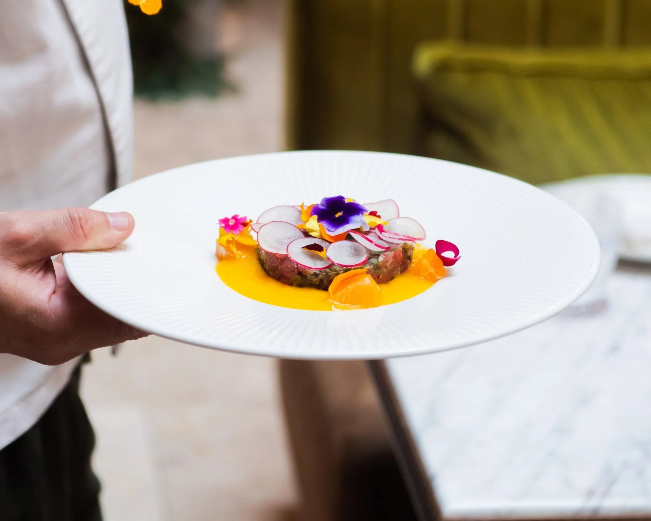 Chef tenant une assiette de tartare dressée avec des tranches de radis et des fleurs comestibles dans un restaurant