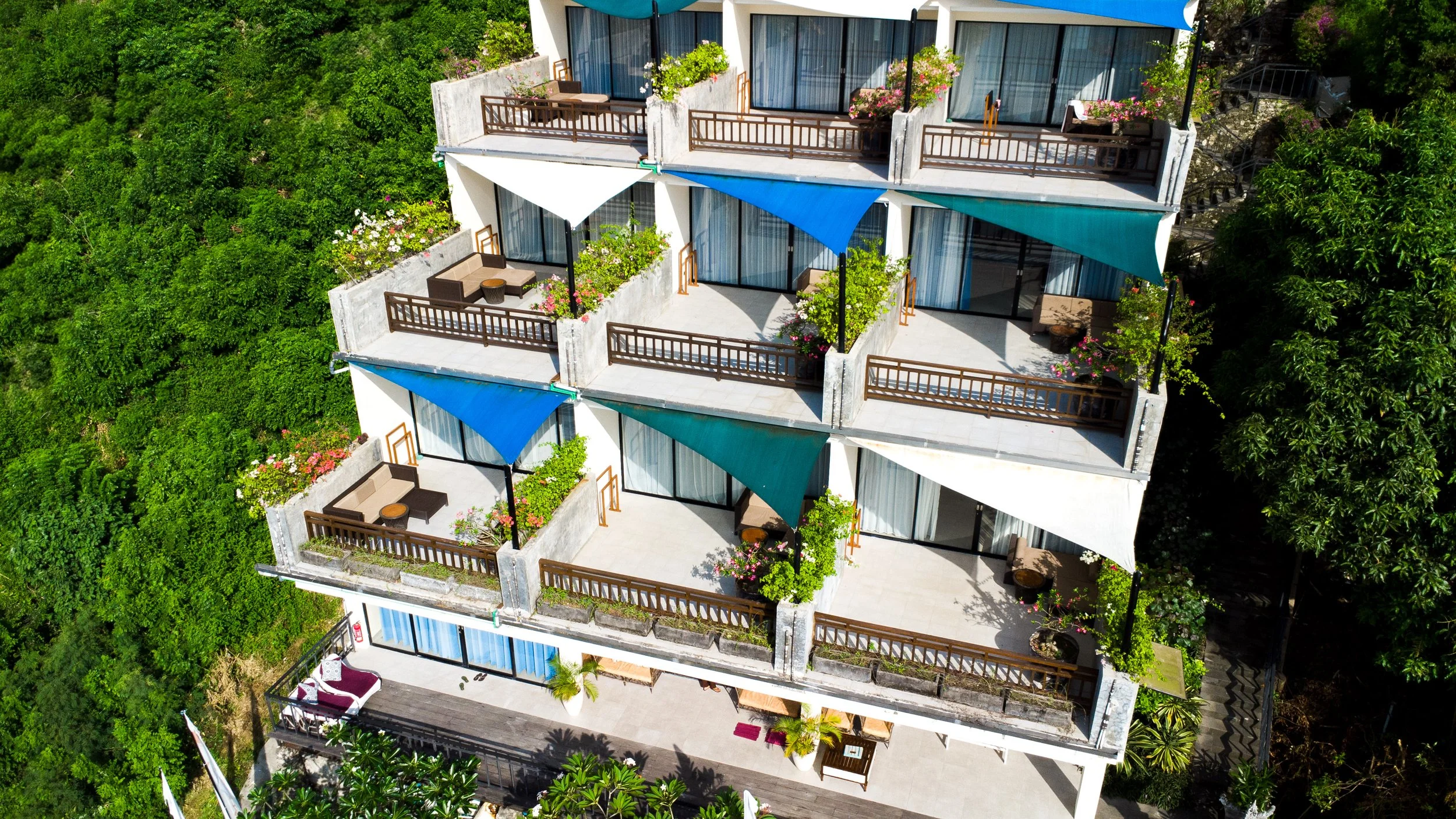 Vue aérienne d'un hôtel avec plusieurs balcons, certains avec parasols bleus ou blancs, des plantes et meubles, entouré de verdure.