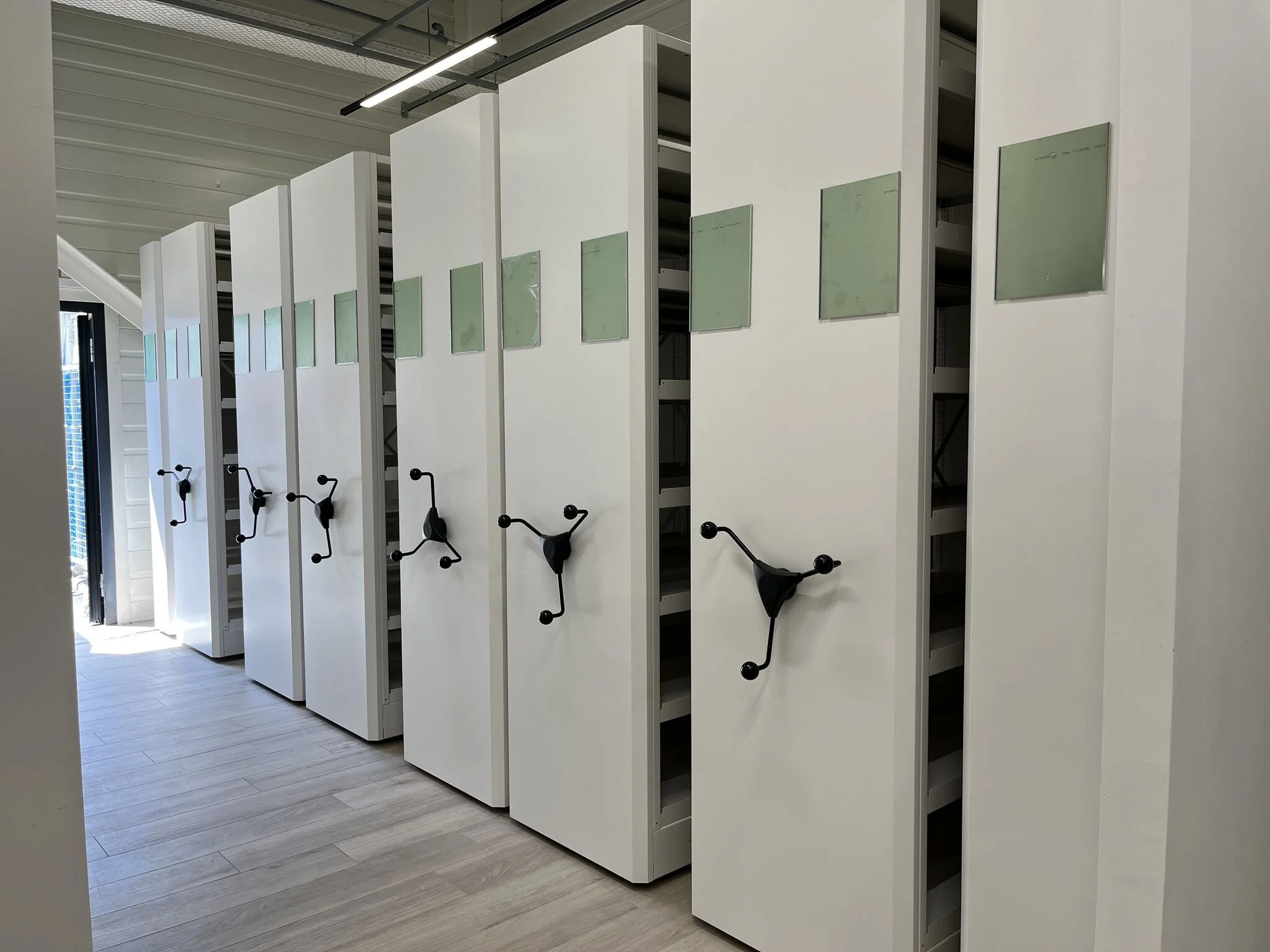 Plusieurs casiers blancs avec des poignées rotatives noires, certains avec des fenêtres de visualisation vertes, dans une pièce avec un plancher en bois.