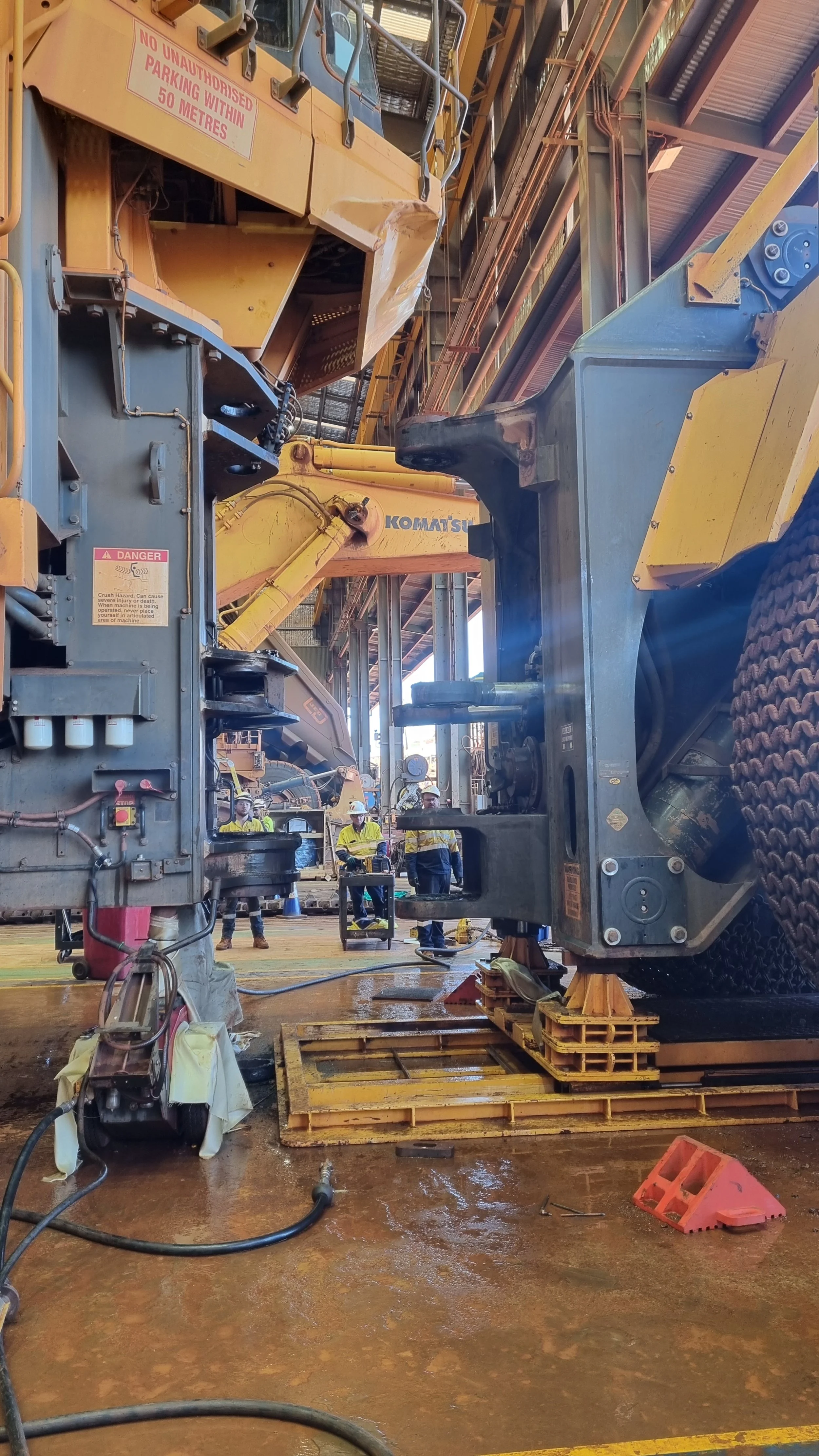 Construction workers in yellow safety vests and white helmets working inside an industrial facility with large machinery, including excavators and metal structures. The floor is wet, with tools, pallets, and safety signs visible.