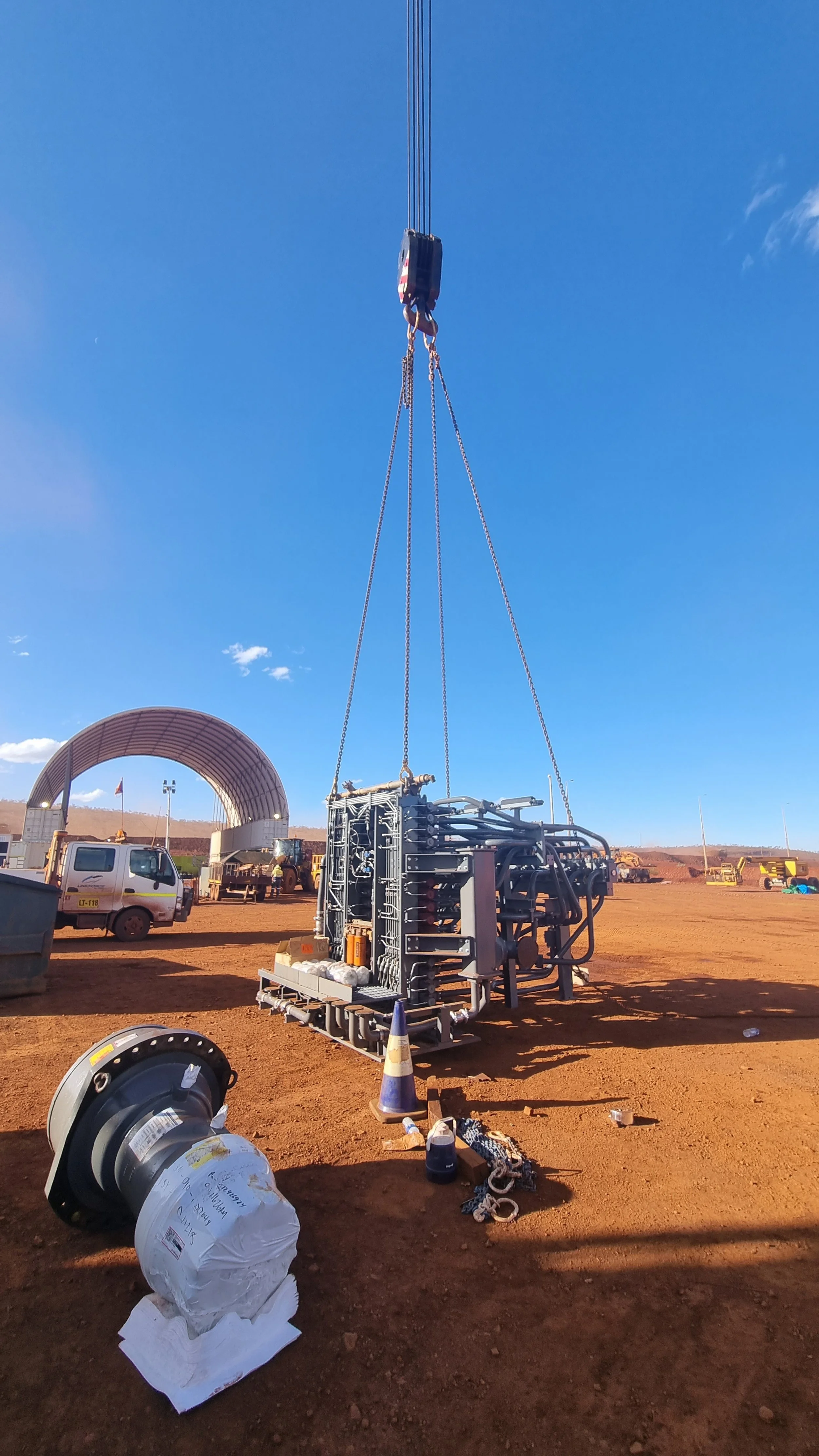 Large piece of industrial equipment being lifted by a crane in an open construction site, with trucks and a large curved shelter in the background under a clear blue sky.