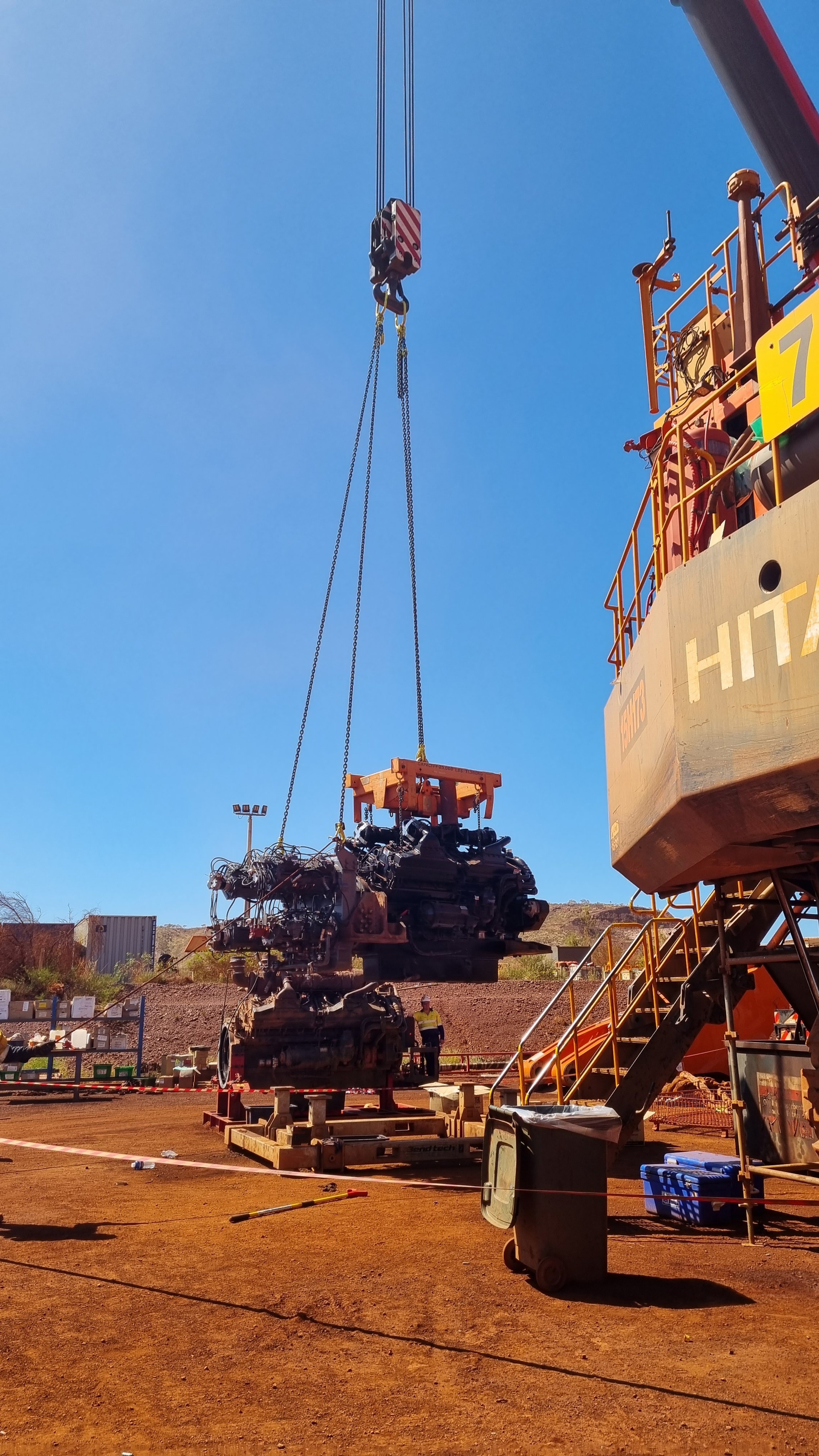 A large crane lifting heavy machinery on a construction site with a dirt ground, a few workers, and a clear blue sky.