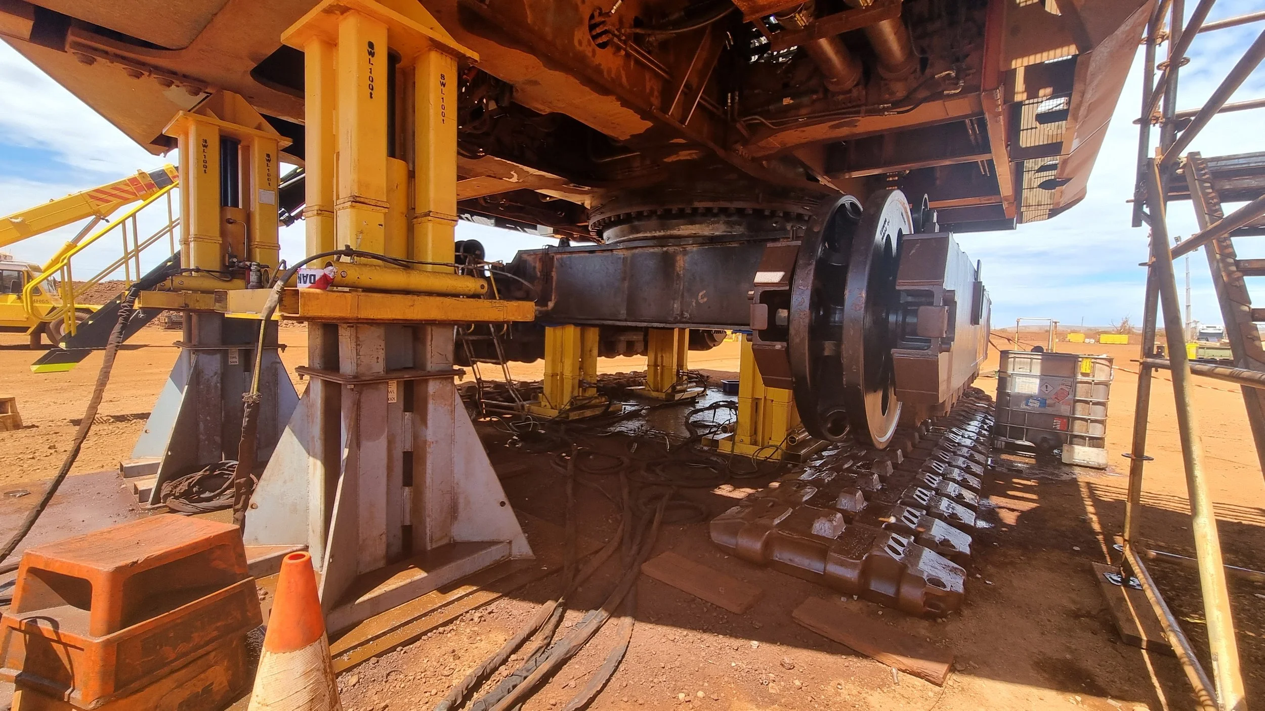 Close-up view of heavy construction machinery with large metal tracks, hydraulic equipment, and a subterranean section, situated on a dirt construction site with a cloudy sky in the background.