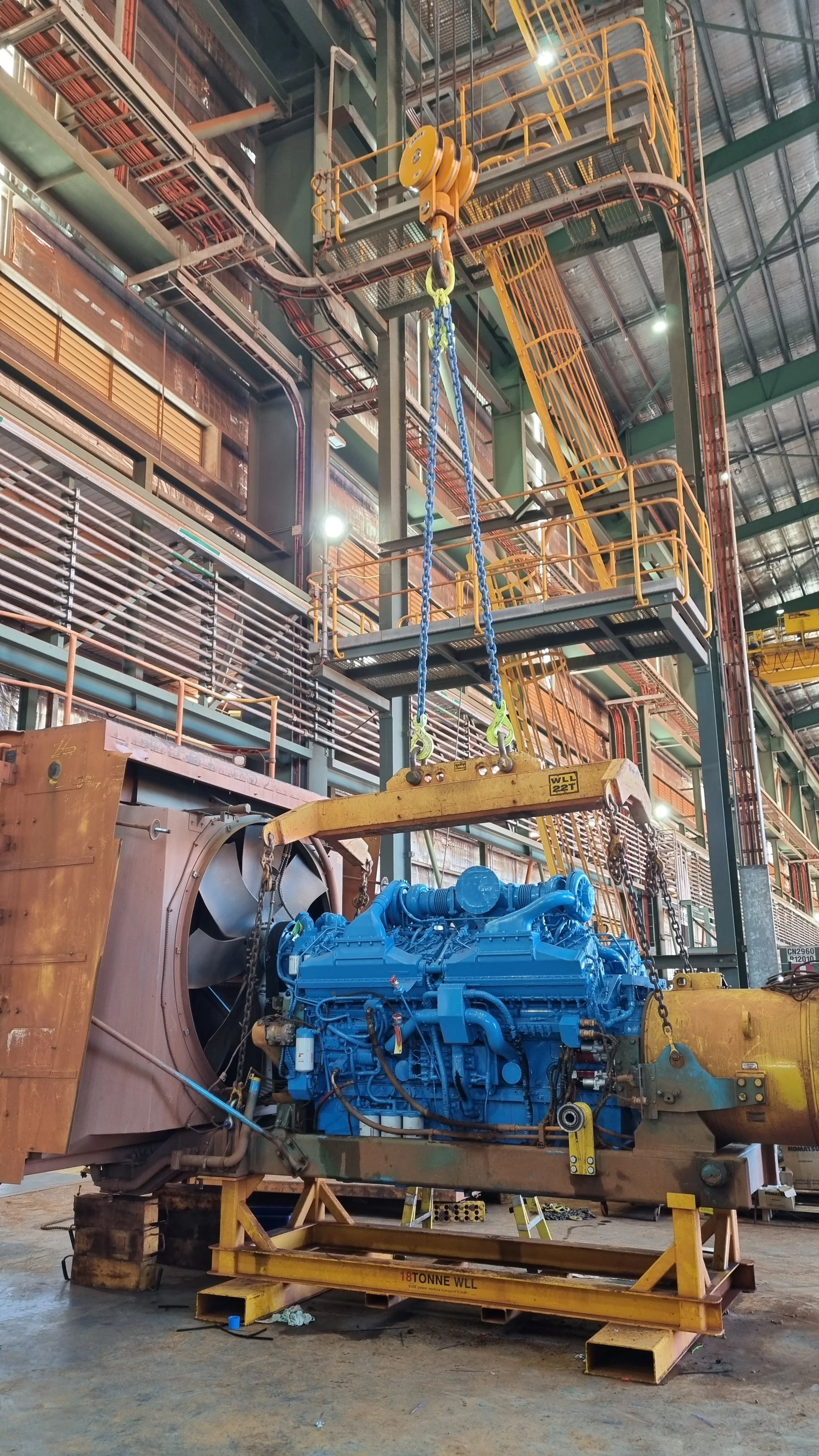 Large blue industrial engine being installed or maintained inside a factory with overhead cranes and metal walkways.