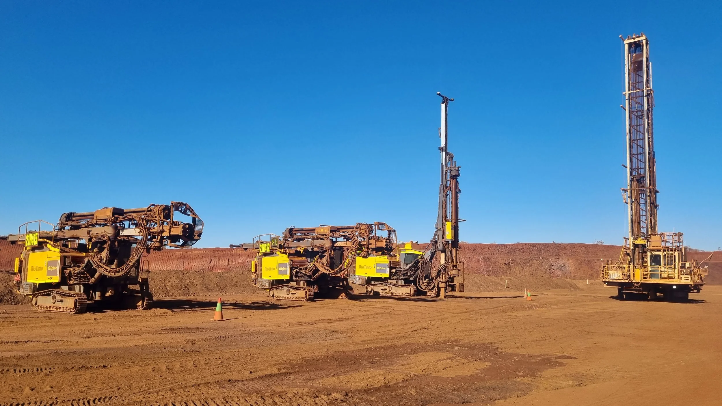 Three large yellow drilling machines on a dirt construction site under a clear blue sky