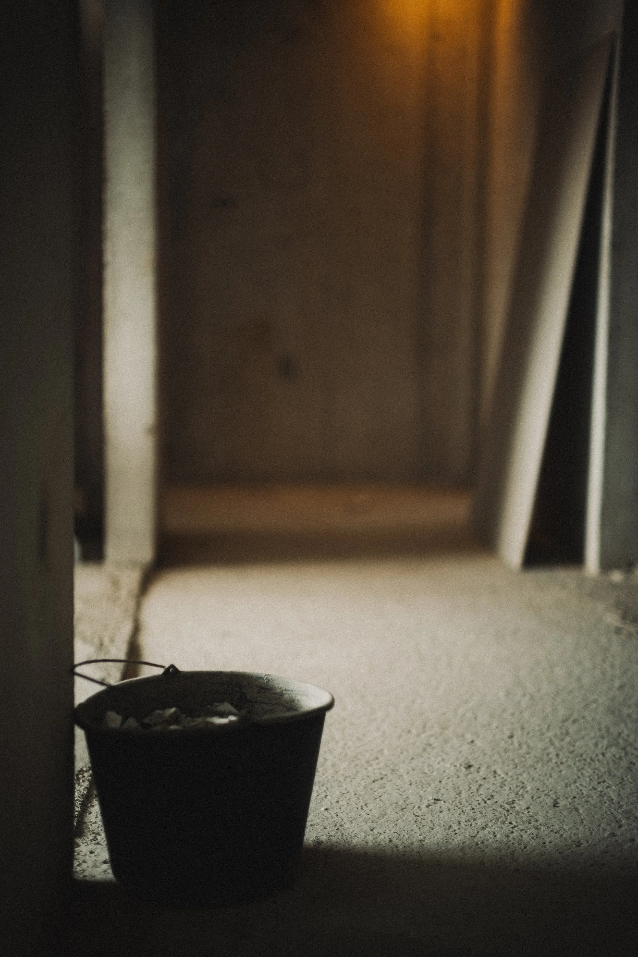 En svart hink med verktyg eller tegelstenar inuti, placerad på ett cementgolv i ett utrymme under byggnation eller renovering, med träskivor och väggar i bakgrunden.