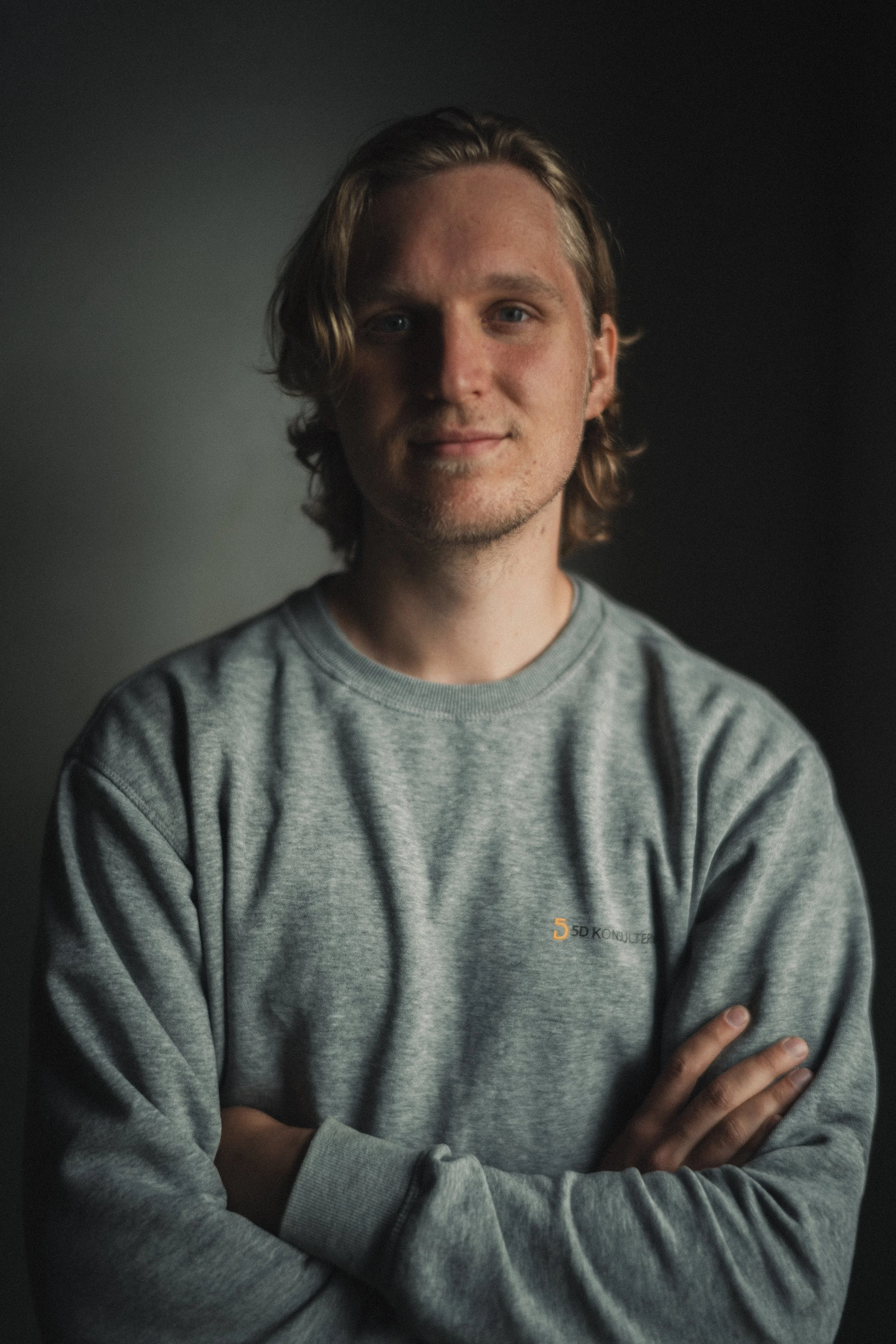 Ung man med blont, vågigt hår, klädd i en grå sweatshirt, står med armar korsade framför en mörk bakgrund.
David Karlsson, Mättekniker.
