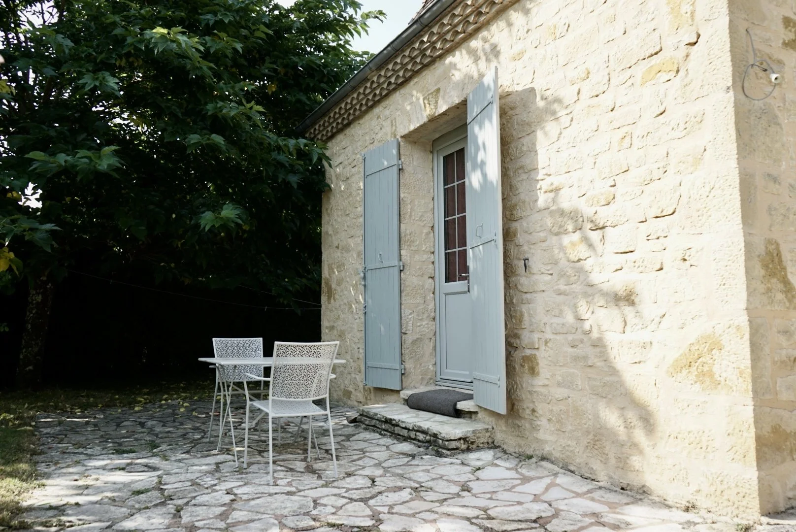 A stone house with light blue shutters, a glass door, and an outdoor patio with a white metal table and chairs, surrounded by greenery.