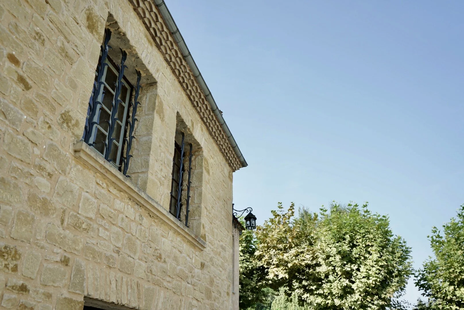 The corner of a stone building with barred windows and a decorative lamp, with trees and a clear blue sky in the background.