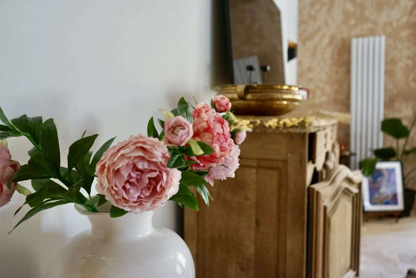 Pink peonies in a white vase with a wooden chest and a framed picture in the background.