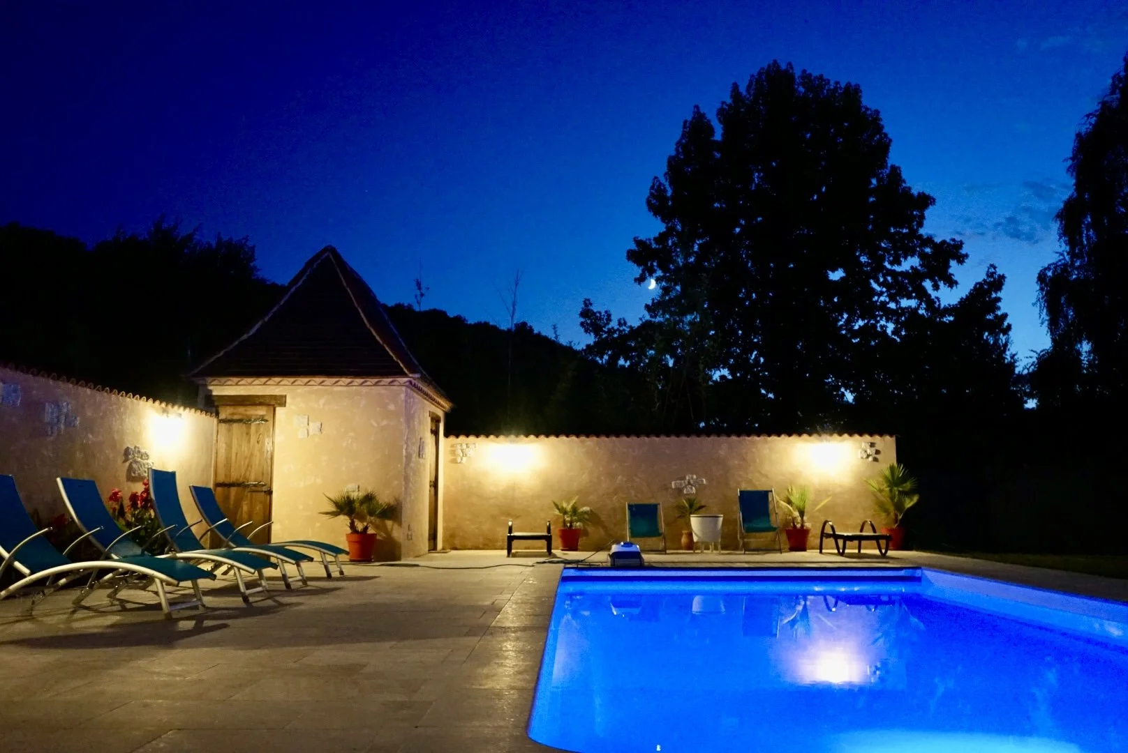 Nighttime scene of a backyard with a lit swimming pool, lounge chairs, potted plants, and a stone wall with lights, surrounded by trees and a dark blue sky.