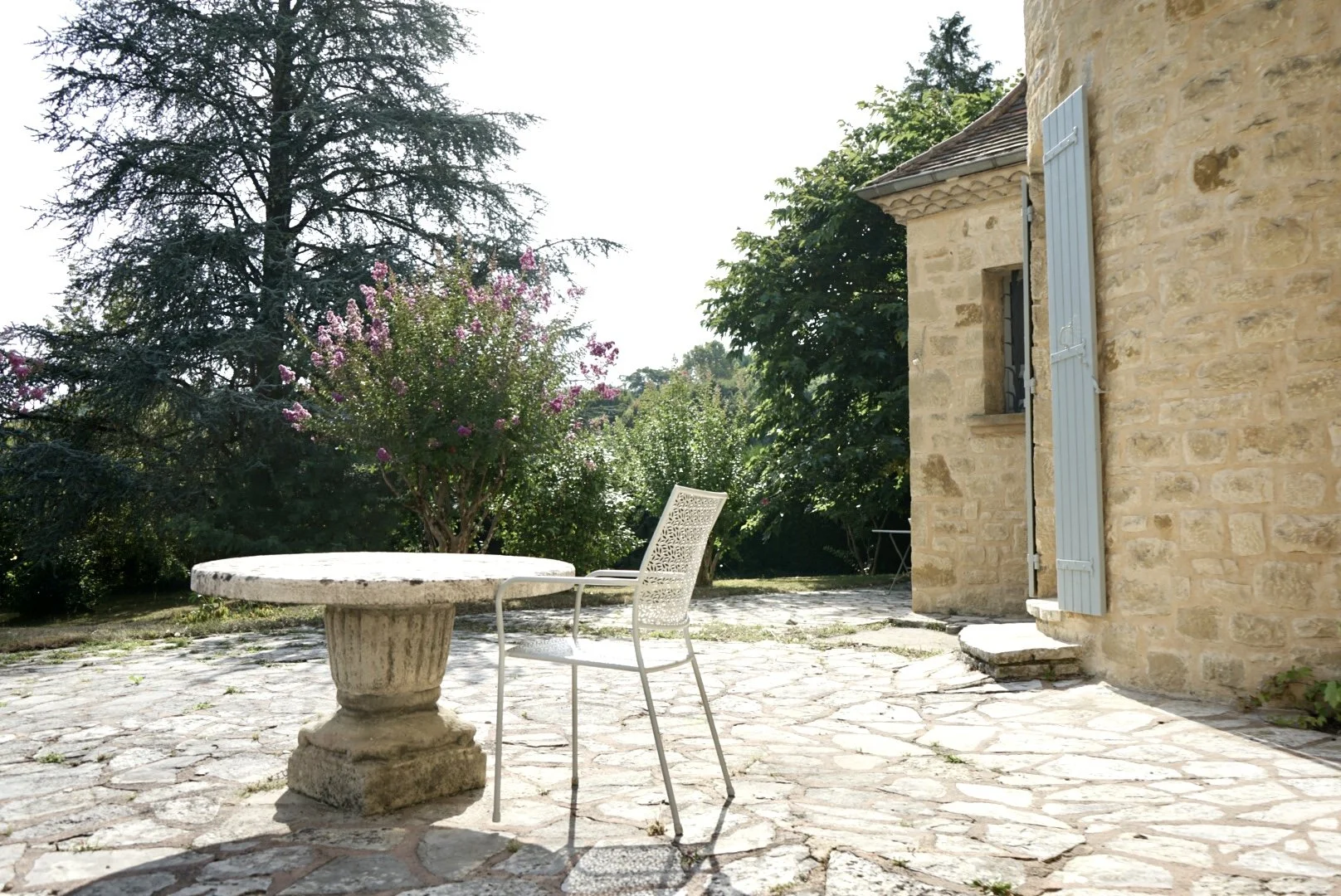 Stone patio outdoor space with a round stone table, a white metal chair, a stone house with blue shutters, and lush green trees and flowering shrub in the background.