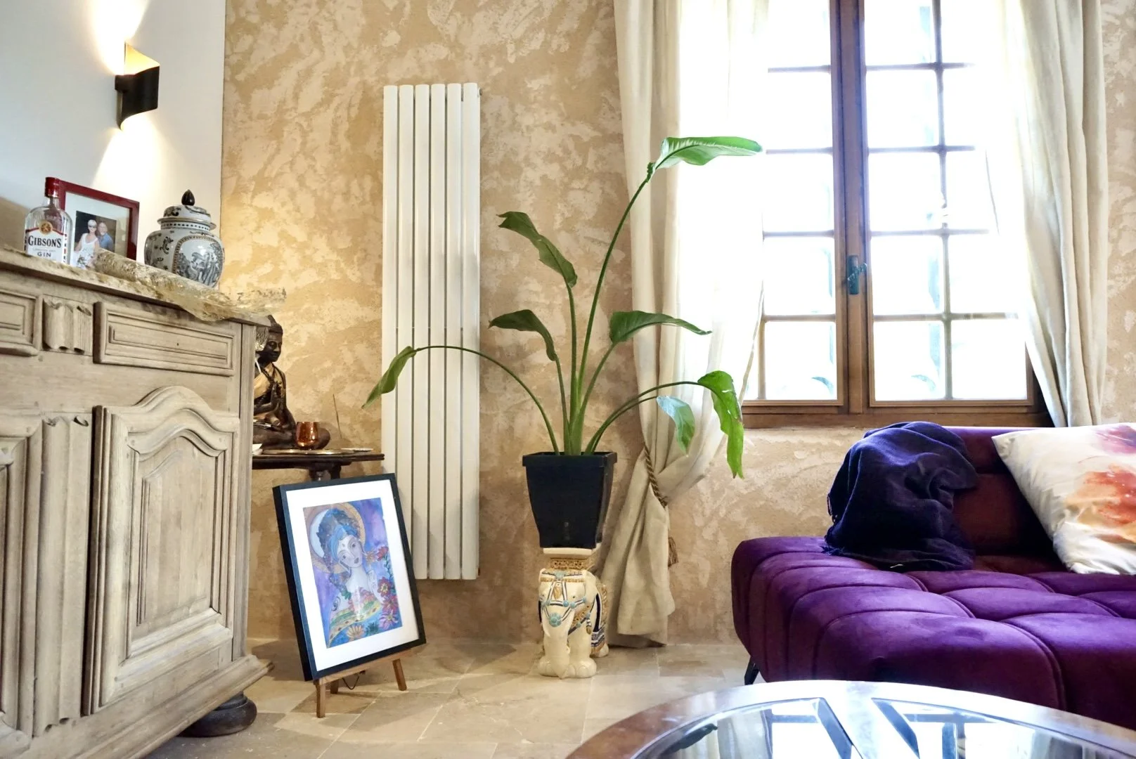Living room with a purple sofa, large window with cream curtains, a large potted plant, decorative items on a wooden sideboard, a framed artwork on the floor, and a radiator on the wall.
