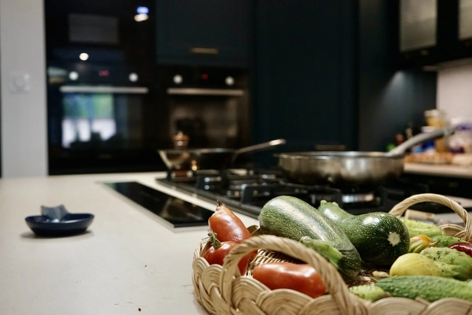Basket of fresh vegetables including zucchini, cucumbers, and tomatoes on kitchen countertop with stove and cookware in background.