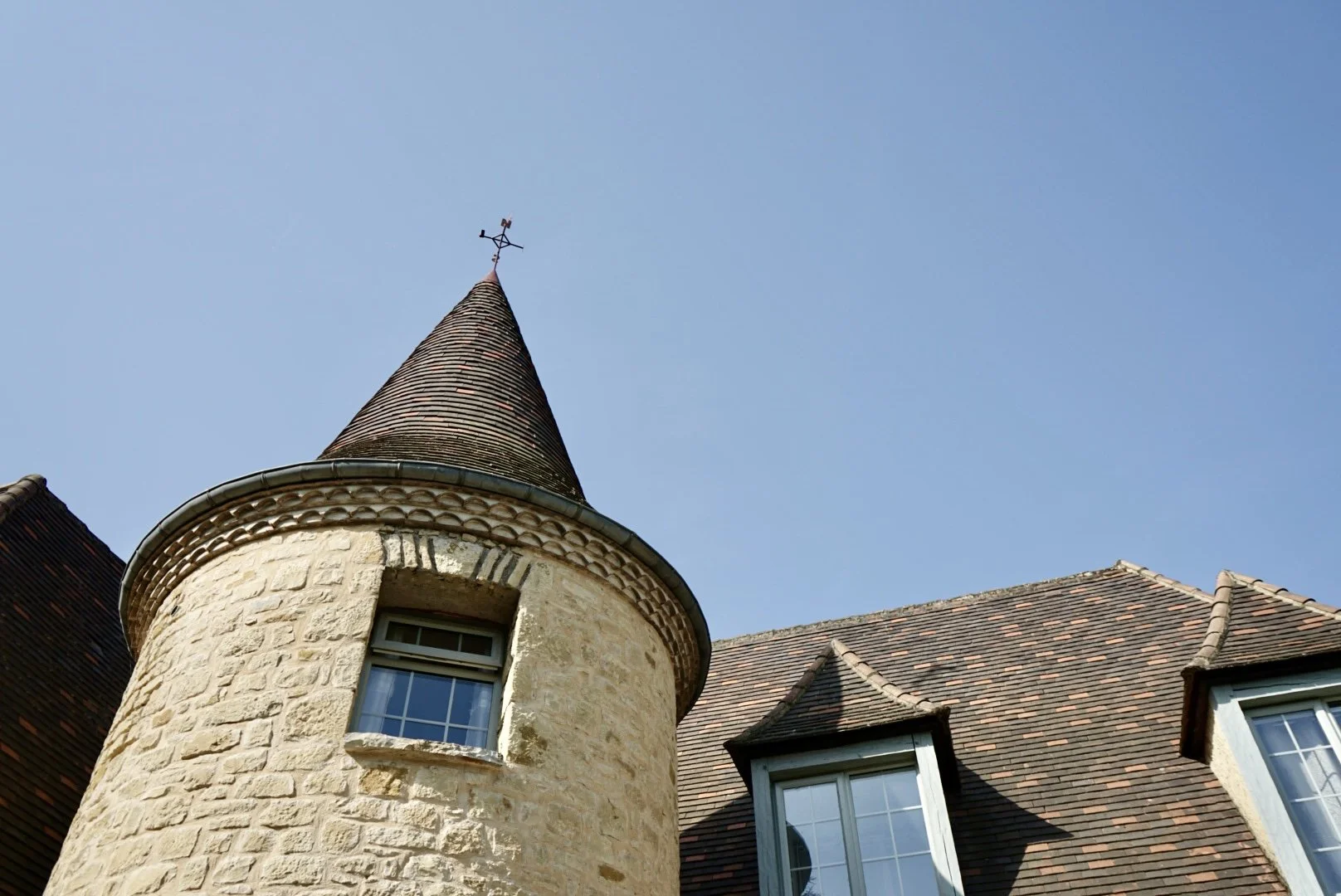 Close-up of a historic stone house with a conical turret and a weathervane on top, set against a clear blue sky.