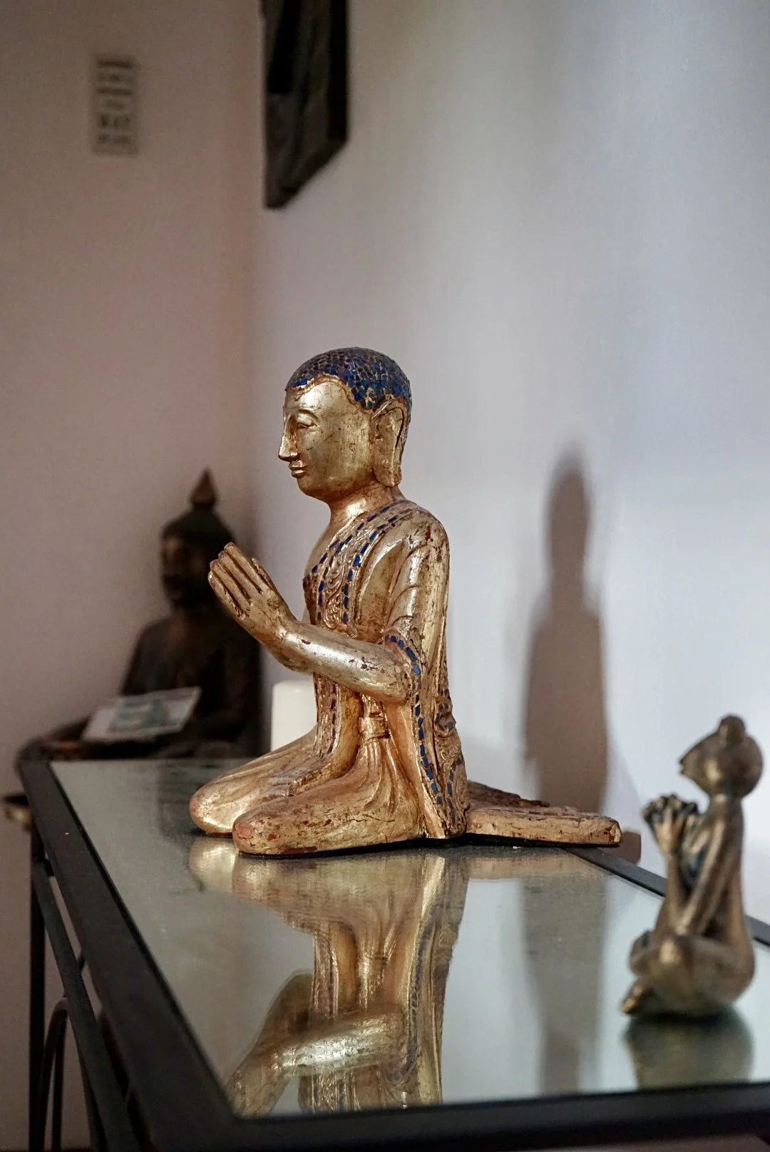 A small, golden, and blue painted Buddha statue in a kneeling position with hands pressed together in a prayer gesture, placed on a glass surface.