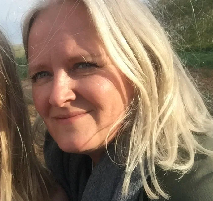 Close-up of a young woman with dark curly hair, wearing glasses, earrings, and layered necklaces, smiling in a bright indoor setting.