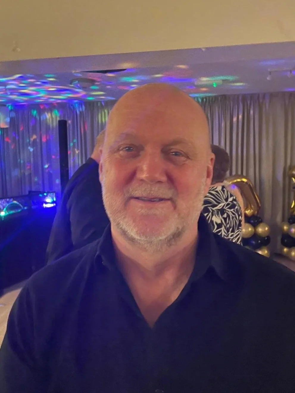 Man in white shirt holding glasses, standing with his back against a white wall, looking downward thoughtfully in an office or home setting with large windows.