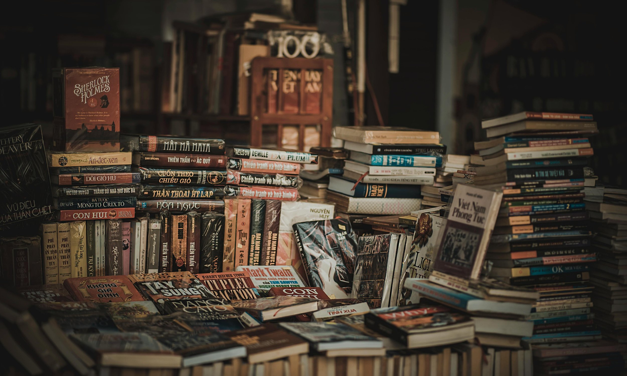 A crowded table filled with various books, with bookshelves in the background.