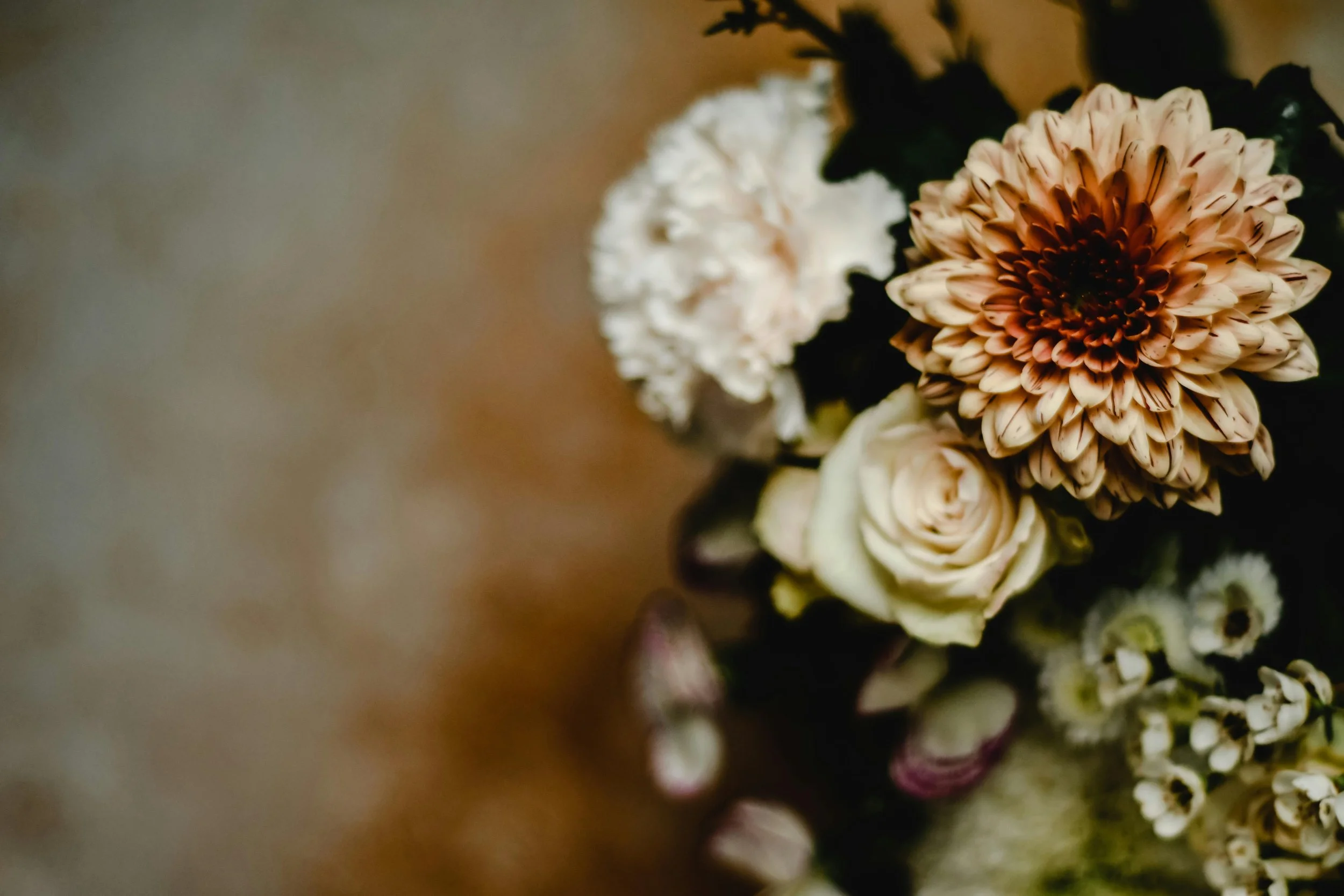 Close-up of a bouquet of flowers including a large peach-colored chrysanthemum, a white ranunculus, and small white flowers with dark centers, on a blurred background.