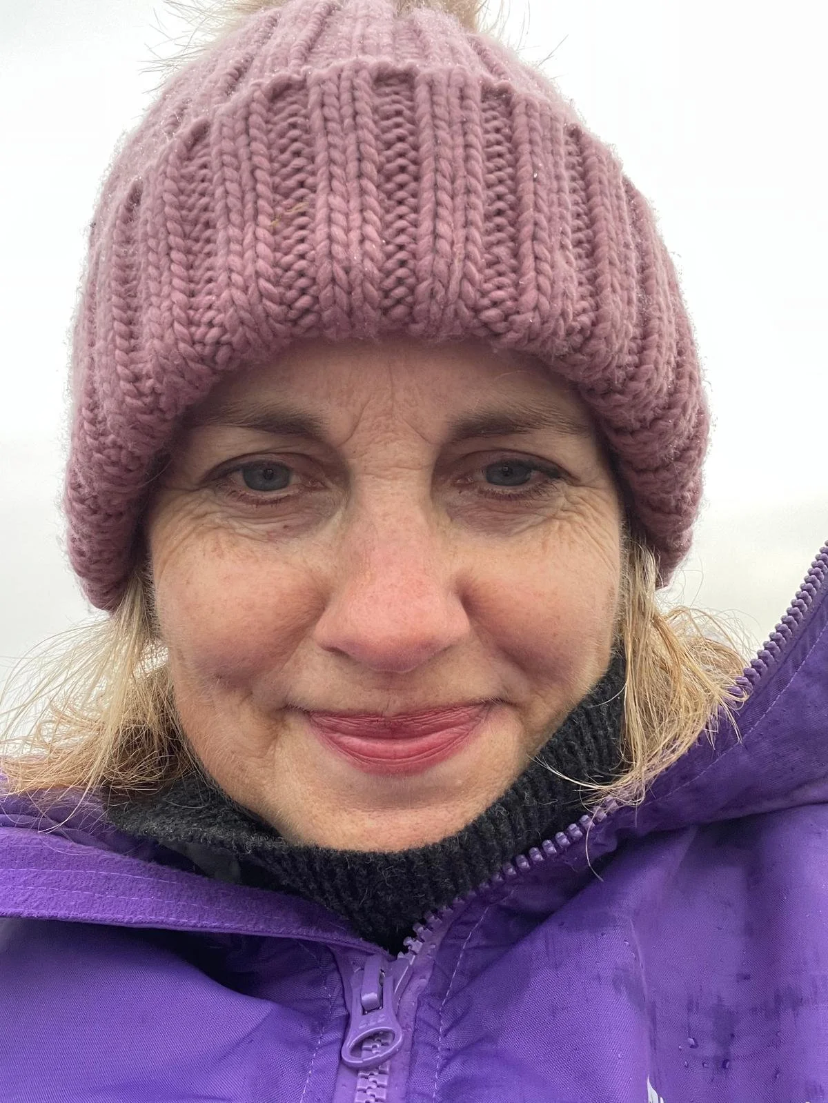 A man with dark, curly hair, wearing clear glasses and a nose ring, looking slightly to the side with a neutral expression, sitting indoors with a blurred plant in the background.