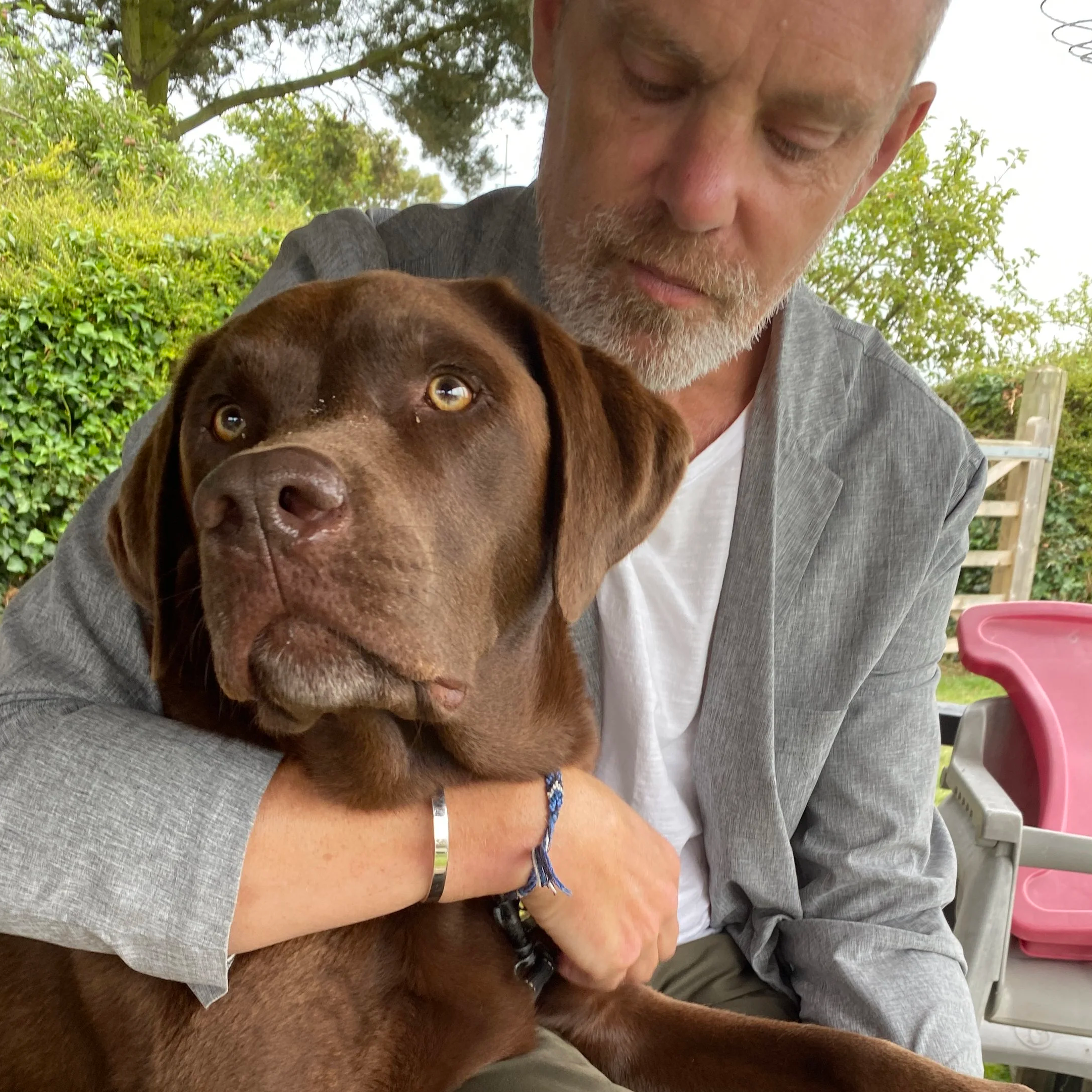 Close-up of a man with curly dark hair, clear glasses, a nose ring, and a beard, wearing a dark green sweater, sitting indoors with a blurred plant in the background.