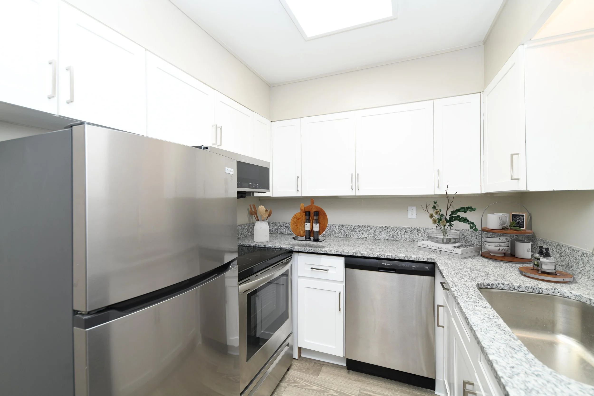 A modern kitchen with white cabinets, granite countertops, stainless steel appliances, and decorative items like a plant, books, and kitchenware.