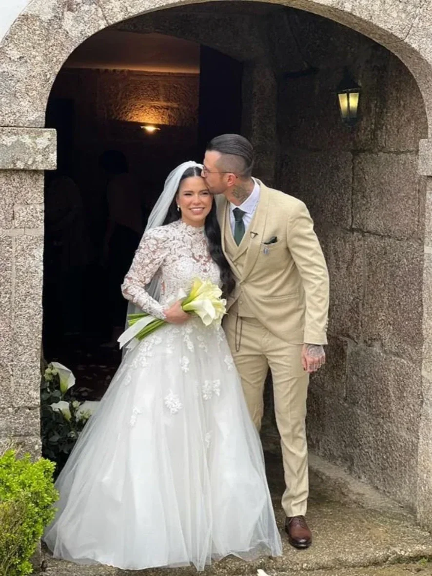 Casal de noivos, ela segura um buquê de flores brancas, ele a beija na testa, ambos sorrindo, na entrada de um lugar com paredes de pedra e iluminação suave ao fundo.