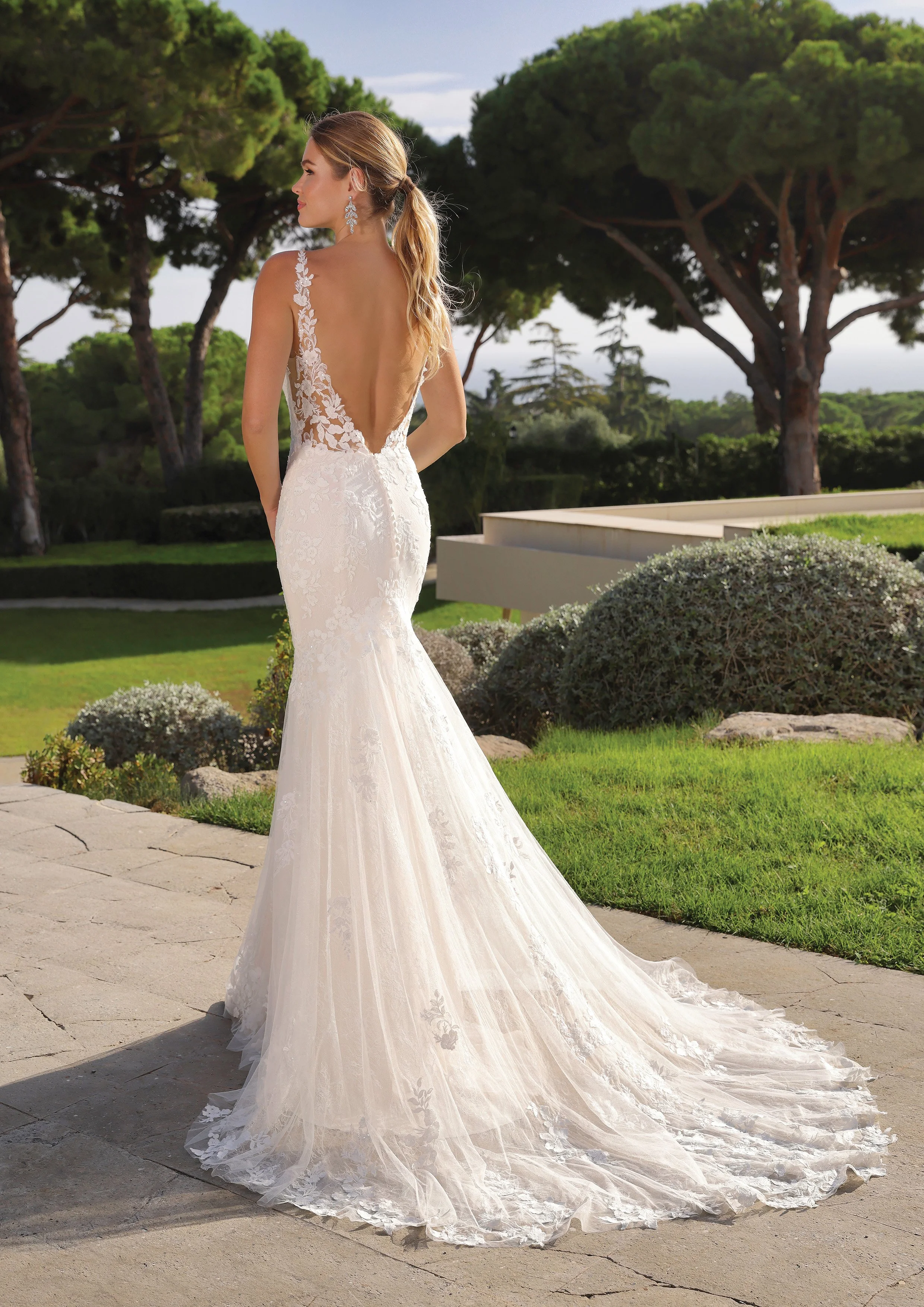 Mulher vestida com vestido de casamento branco, com detalhes de renda floral, vista de costas, ao ar livre, com árvores verdes ao fundo.