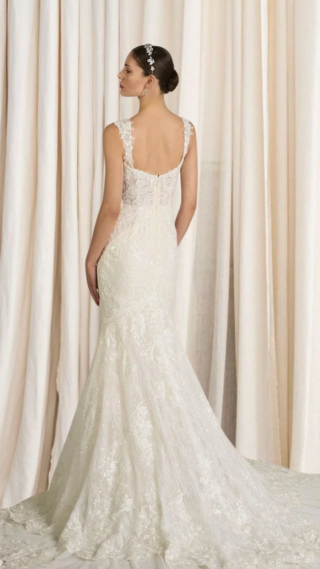 Noiva de vestido de noiva branco com detalhes em renda, de costas, com cabelo preso e acessório floral, em fundo de cortinas brancas.