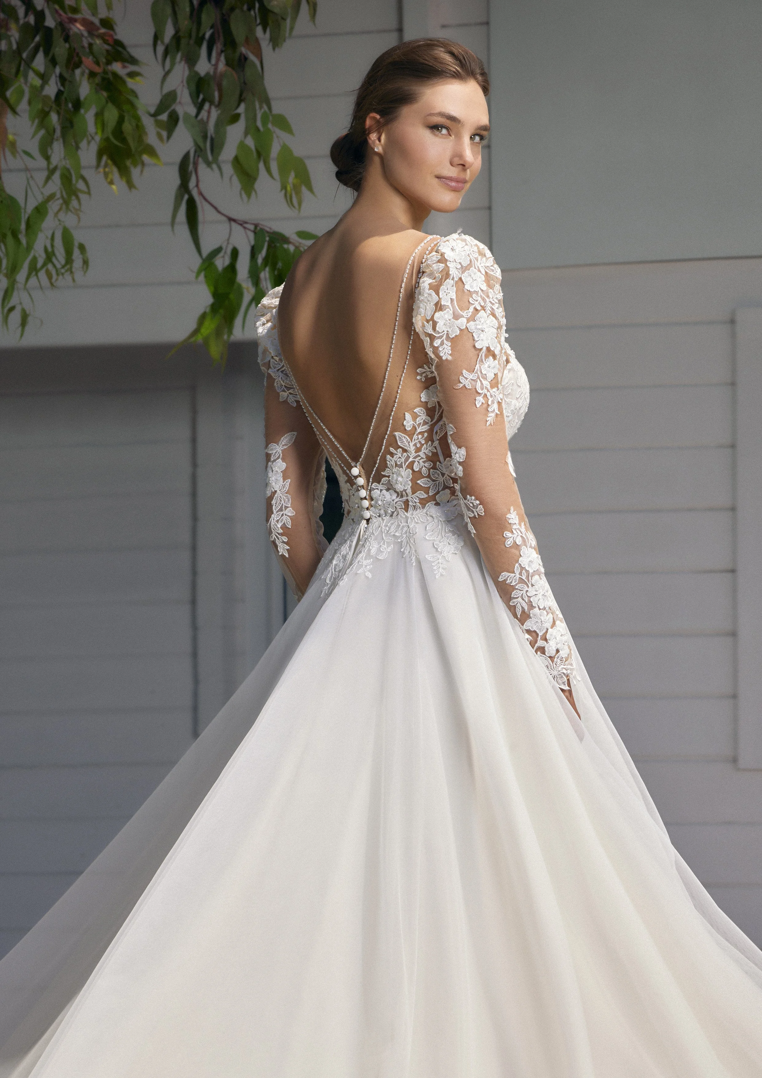 Mulher vestida com vestido de casamento branco, de costas, com detalhes em renda floral e cabelo preso, ao lado de uma árvore verde, em ambiente externo.