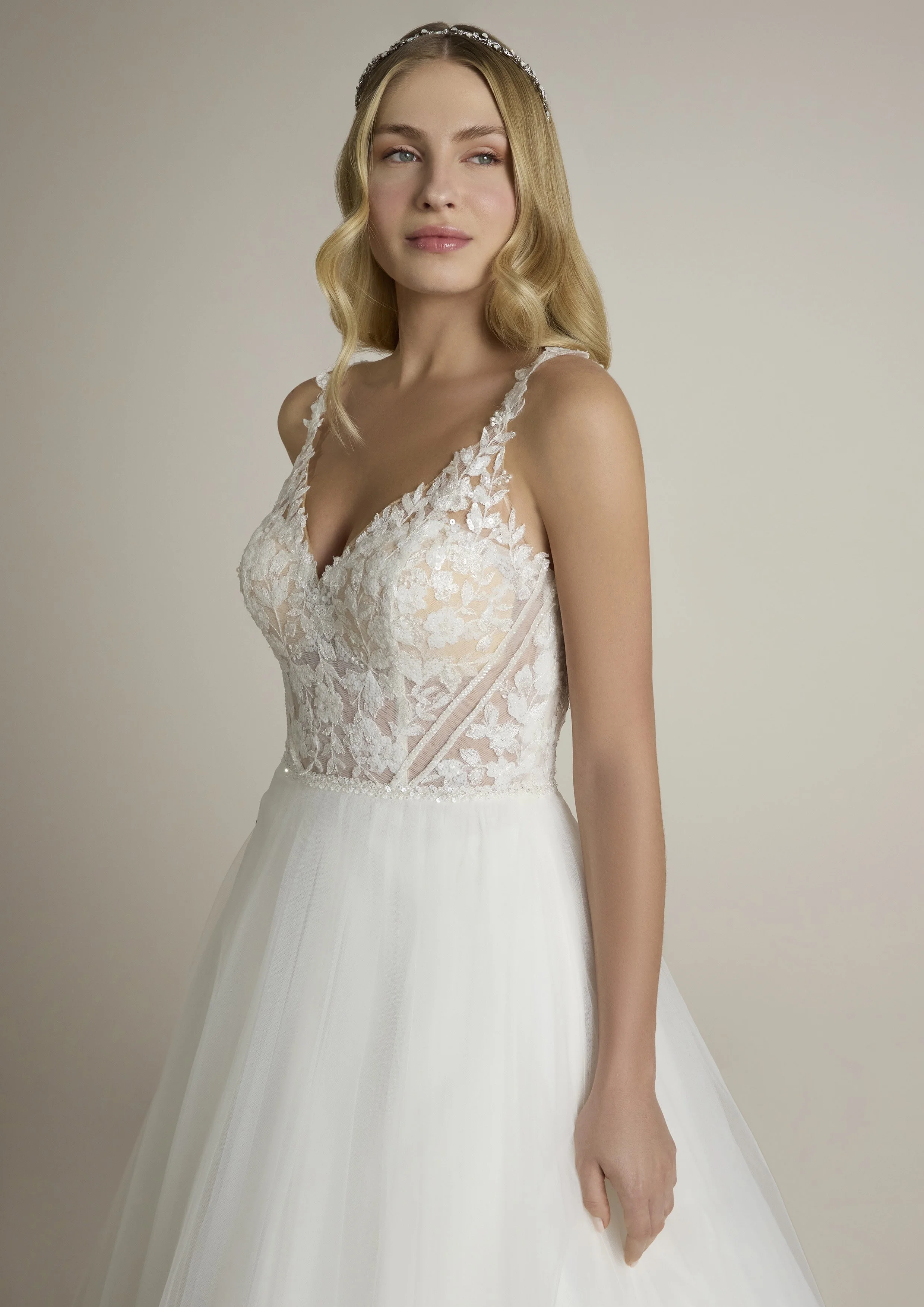 Mulher com cabelo loiro, usando vestido de noiva com decote em V e detalhes de renda floral na parte superior, penteado com ondas suaves, usando diadema de pedras, fundo neutro.