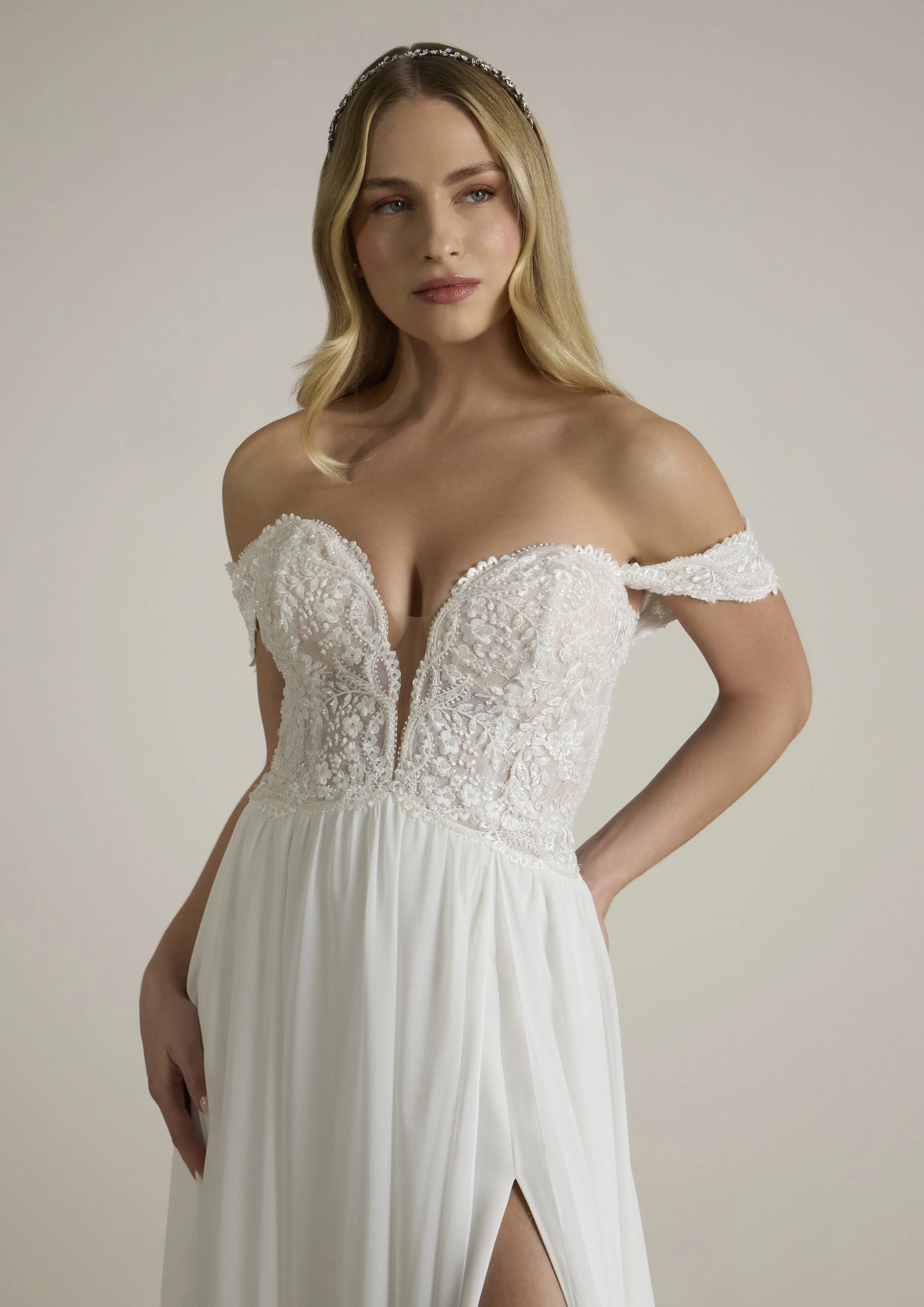Mulher com cabelo loiro ondulado, usando tiara de pérolas, vestido de noiva com detalhes de renda e decote em V, posando contra fundo neutro.