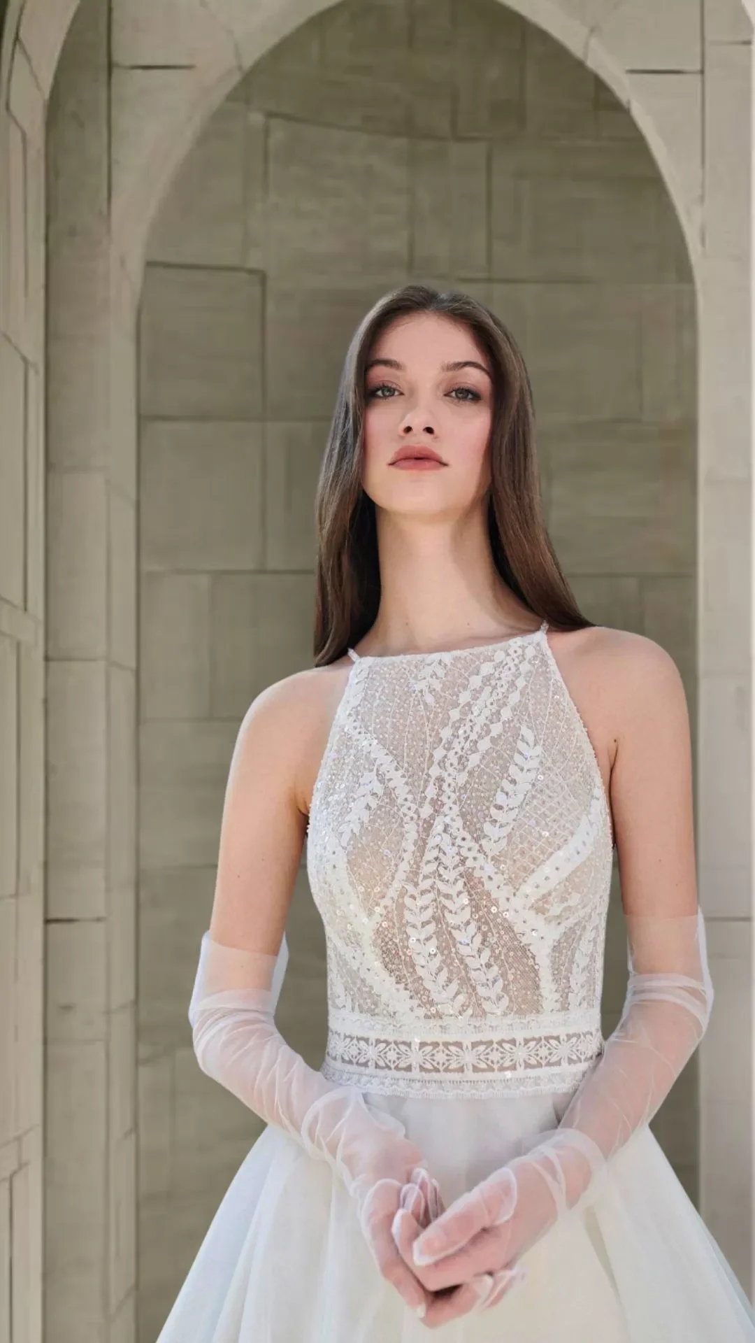 Mulher com cabelo castanho lisos usando vestido de noiva com detalhes em renda e luvas transparentes, em ambiente com paredes de pedra