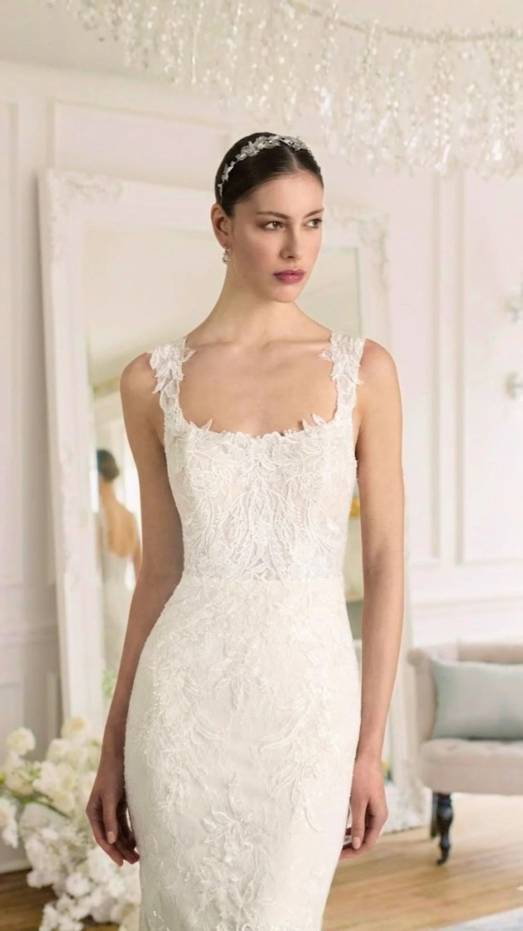 Mulher em vestido de casamento branco com detalhes em renda, usando uma tiara, em um ambiente decorado com flores brancas e espelho ao fundo.