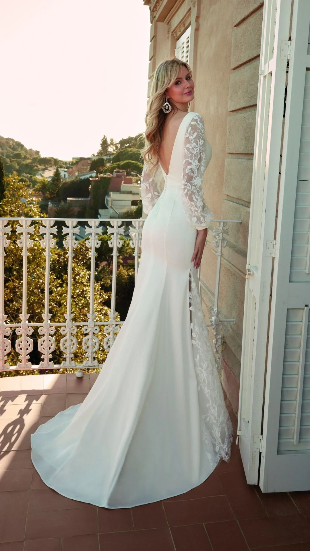Mulher em vestido de noiva branco com detalhes de renda nas mangas, posando em um balcão com vista para árvores e casas ao fundo.