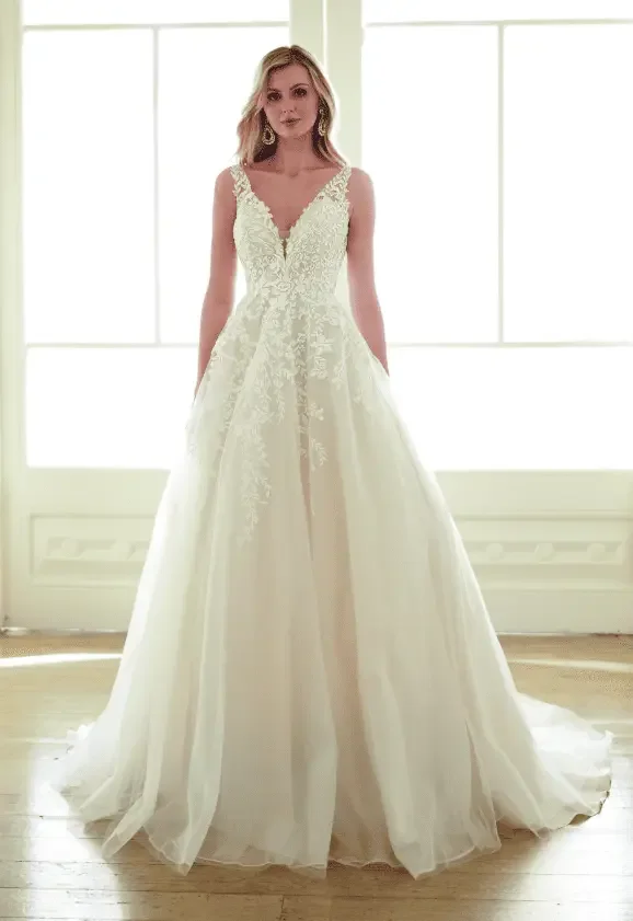 Mulher de vestido de noiva com saia longa e detalhes em renda, de cabelo loiro, com brincos grandes, posando diante de janela com luz forte.