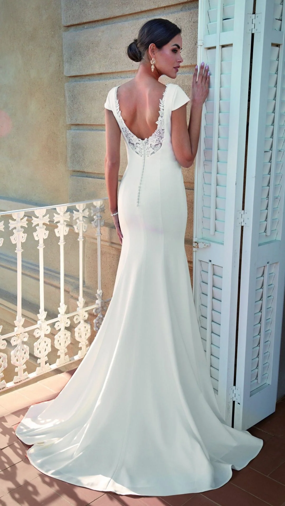 Mulher com vestido de noiva branco, de cauda longa, segurando uma persiana branca na varanda.