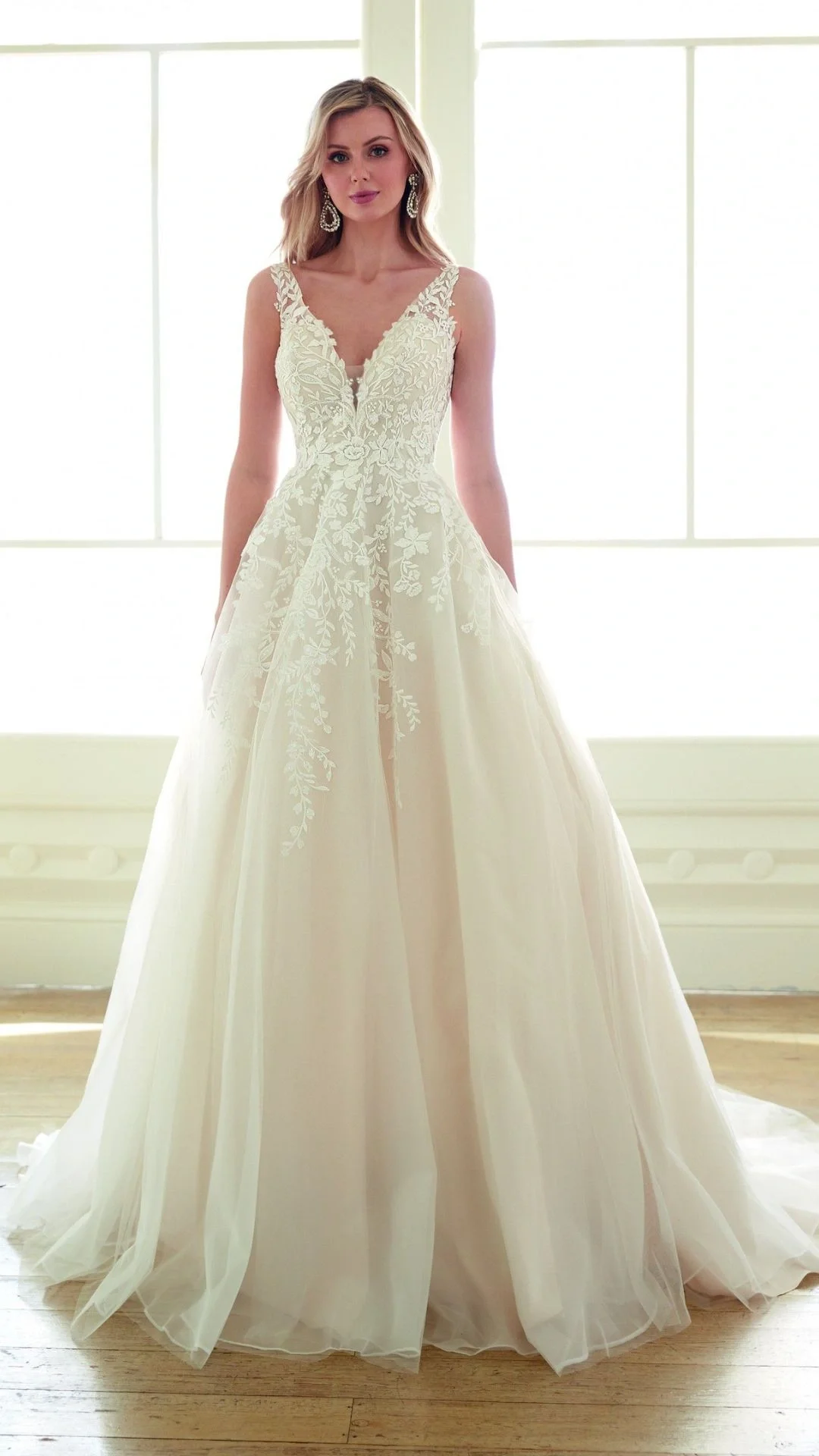 Mulher com vestido de noiva de saia longa em tom off-white, detalhes em renda floral na parte superior, com decote em V, dentro de casa com janelas grandes ao fundo.