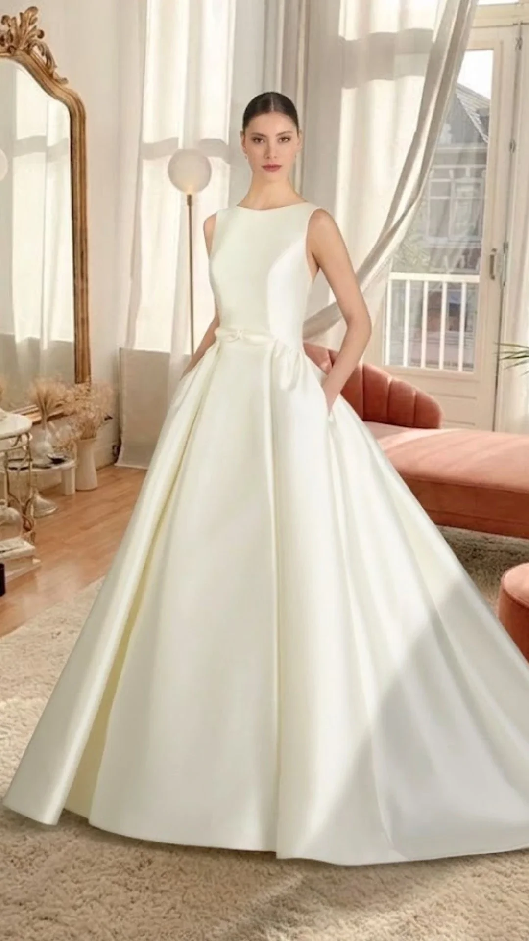 Mulher vestida com vestido de noiva branco, de vestido longo e saia ampla, posando em sala com cortinas brancas, espelho dourado e sofá.